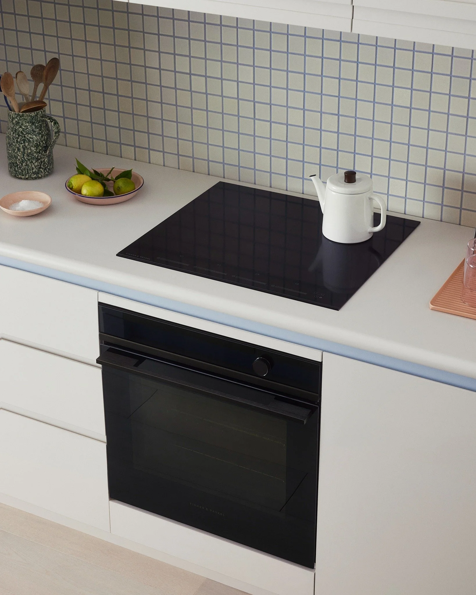 Kitchen countertop with a white electric kettle on a black stovetop, a pink plate with green apples, a small pink dish, a container with wooden utensils, and a pink tray with glassware, with a tiled backsplash.