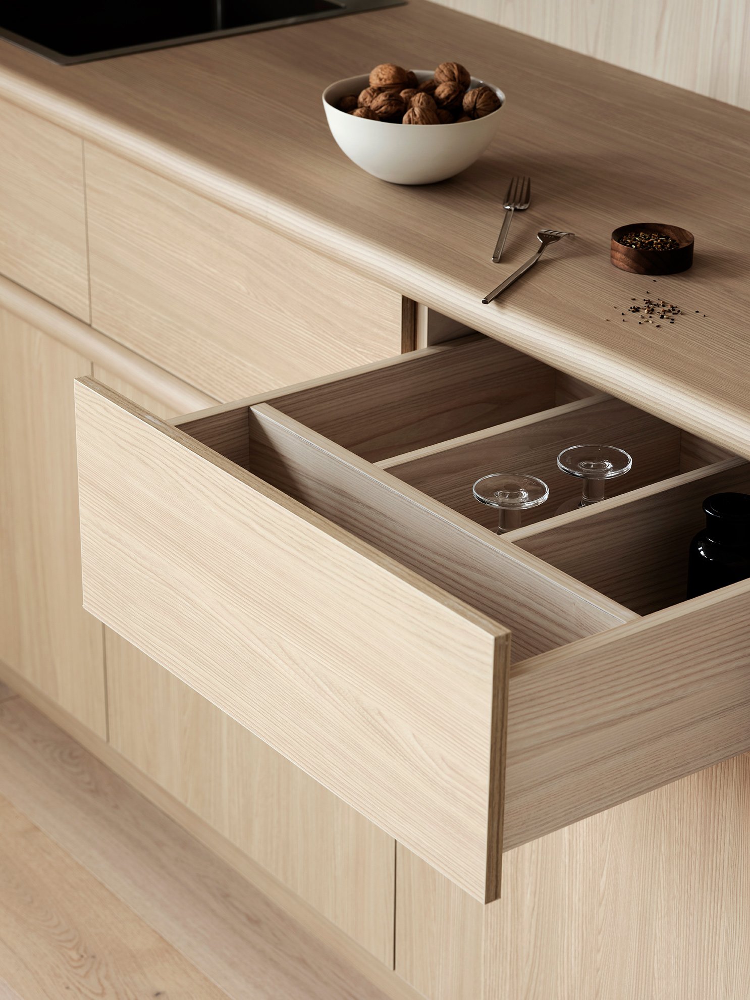 Open wooden drawer with glassware inside, on a wooden kitchen countertop with a bowl of walnuts, a fork, a spoon, and a small bowl of peppercorns on top.