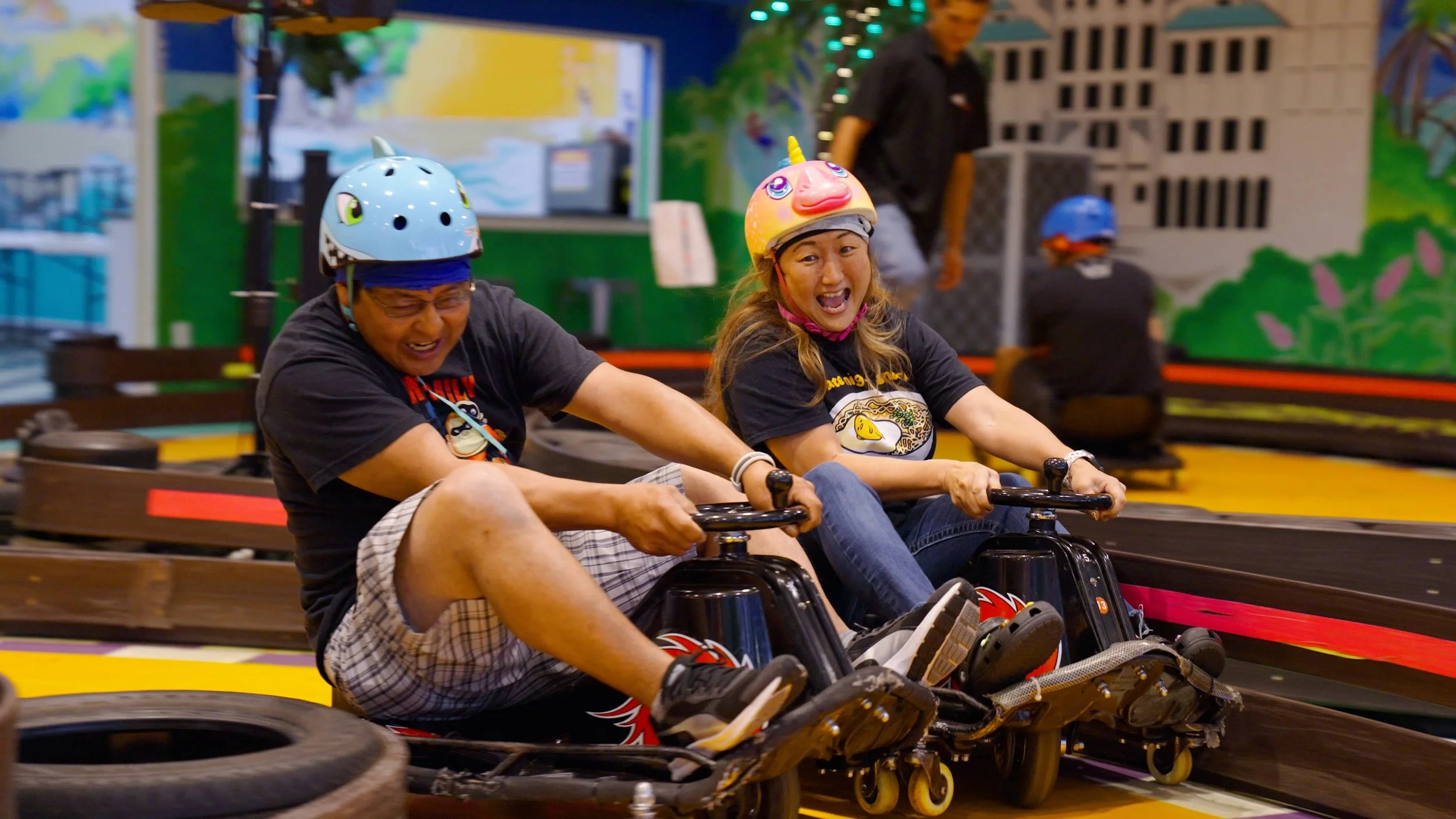 Two people are riding go-karts indoors, both wearing helmets and smiling, with a colorful mural and racing track in the background.