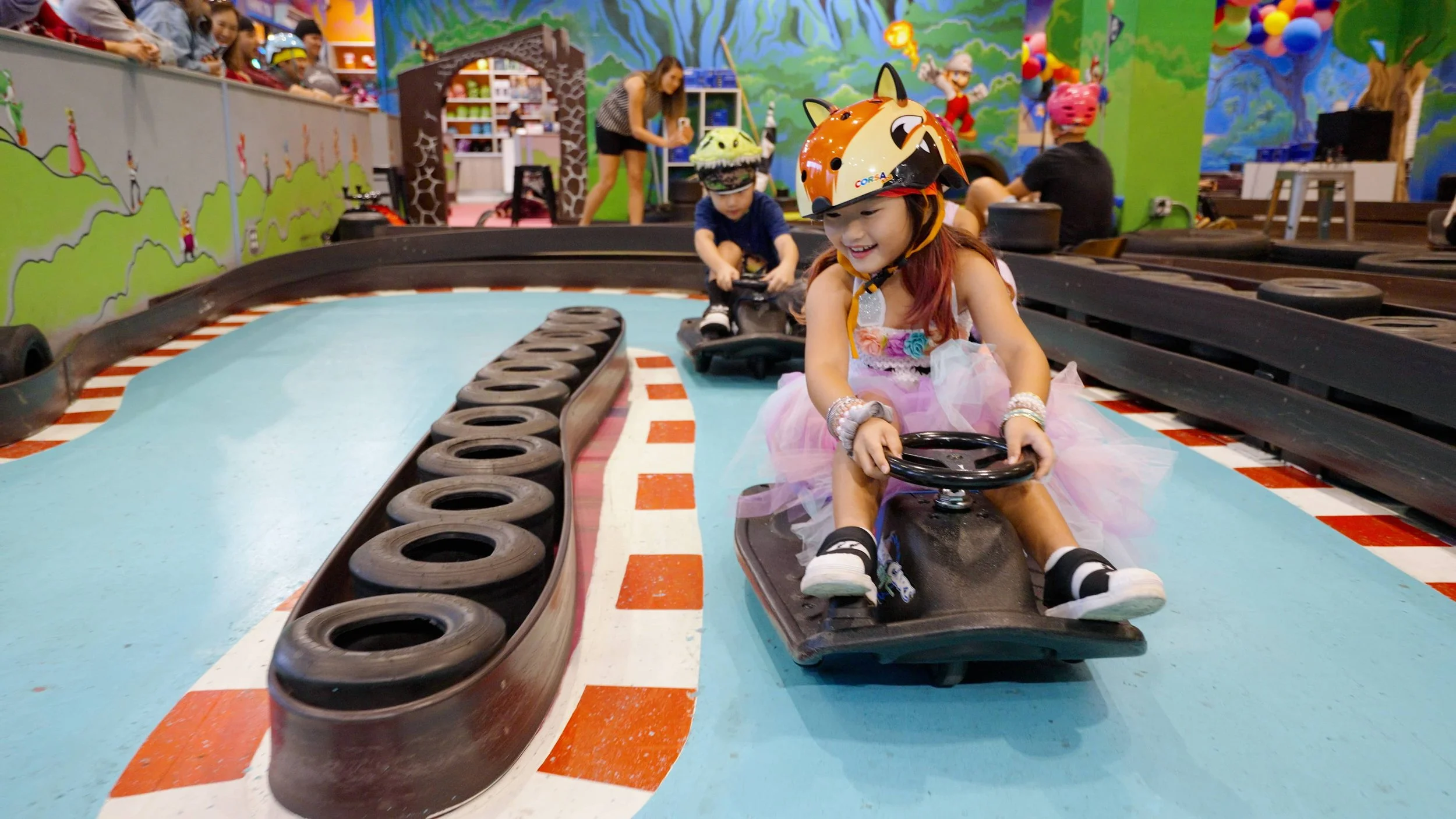 Children driving custom go-karts on an indoor track at an amusement center, with colorful walls and onlookers watching.