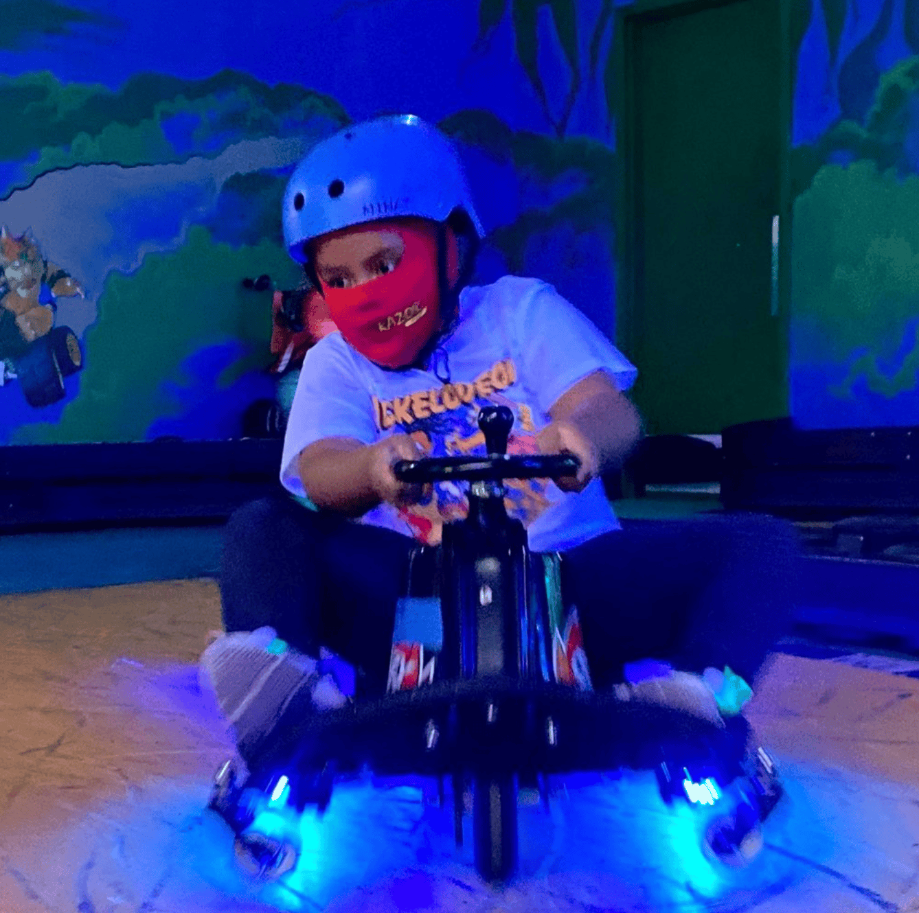Child wearing a helmet and a red face mask riding an electric go-kart with blue lights, in an indoor track with animated forest mural background in a cosmic setting.
