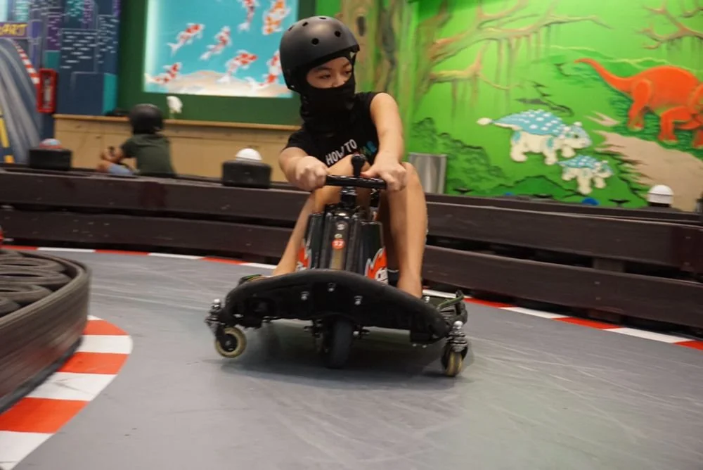A young boy wearing a black helmet and face mask drifting a drift-kart on an indoor race track with colorful murals of animals and nature in the background.
