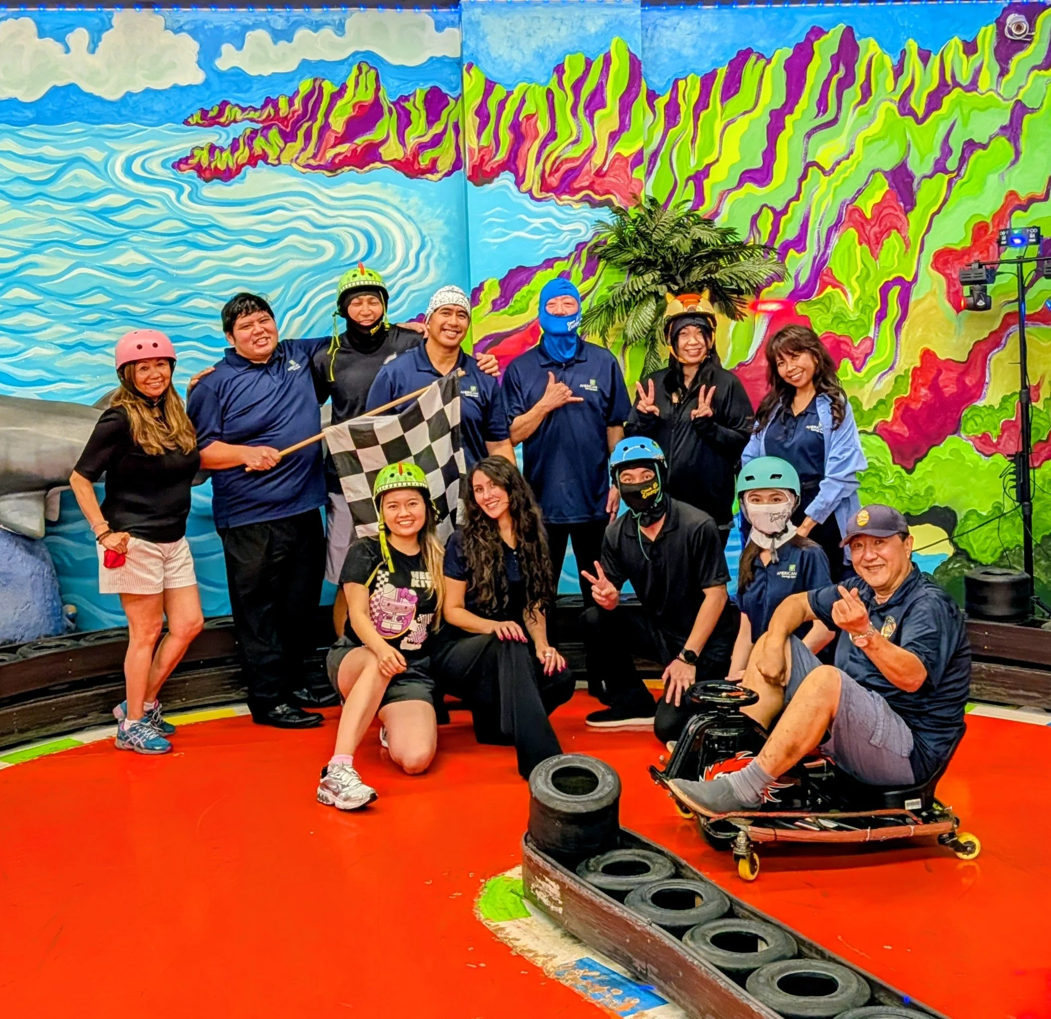 A group of people with helmets and safety gear standing in front of a colorful mural of mountains and water. The floor has red, green, and blue painted sections, and there are tires and a skateboard nearby.