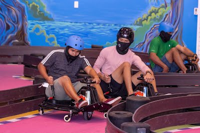 Three people driving drift karts on an indoor track, wearing helmets and casual clothing, with a colorful painted backdrop of trees and water.