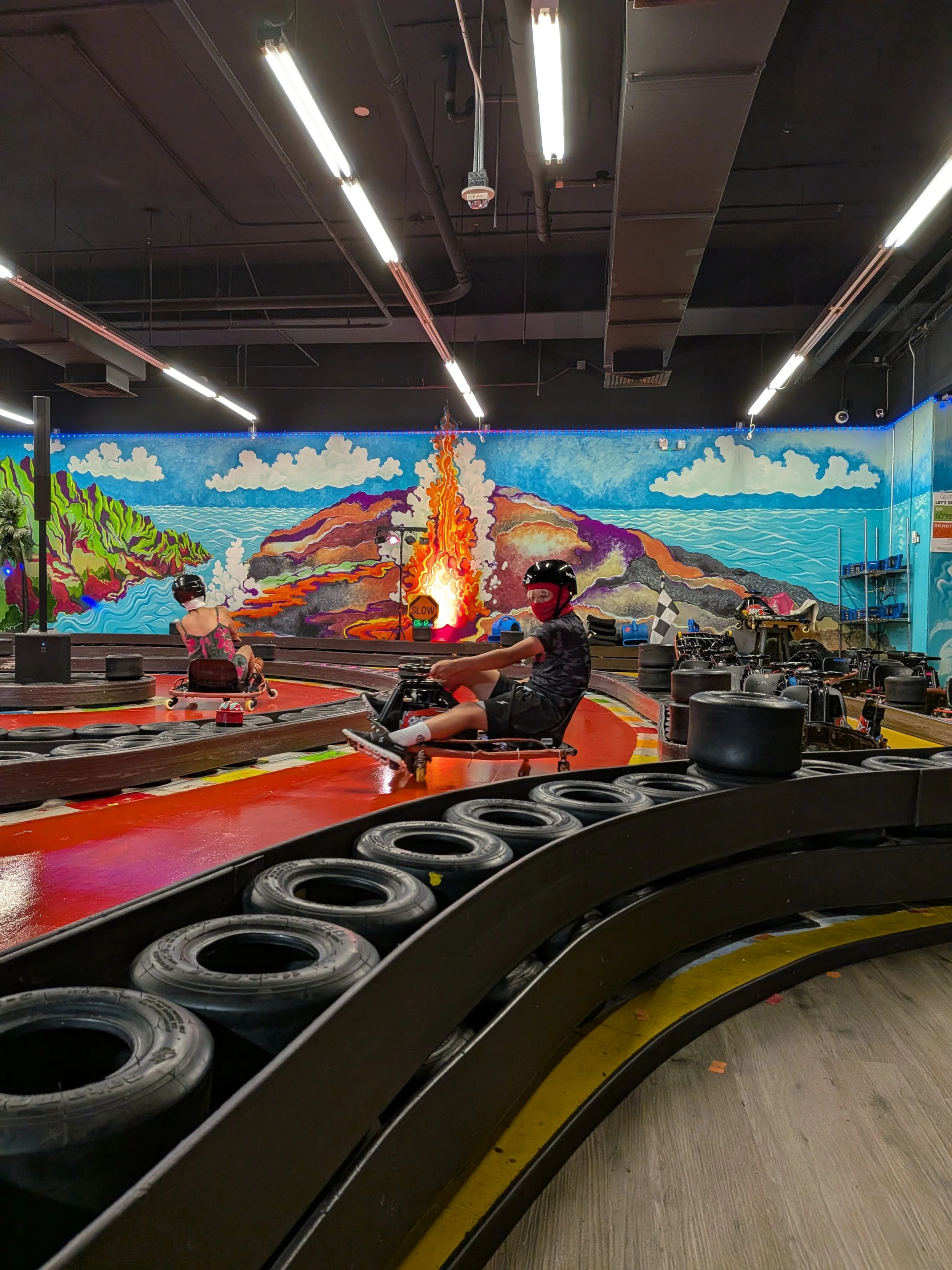 Indoor go-kart racing track with a colorful mountain and sky mural, two children wearing helmets sitting in go-karts, surrounded by tires on the track boundary.