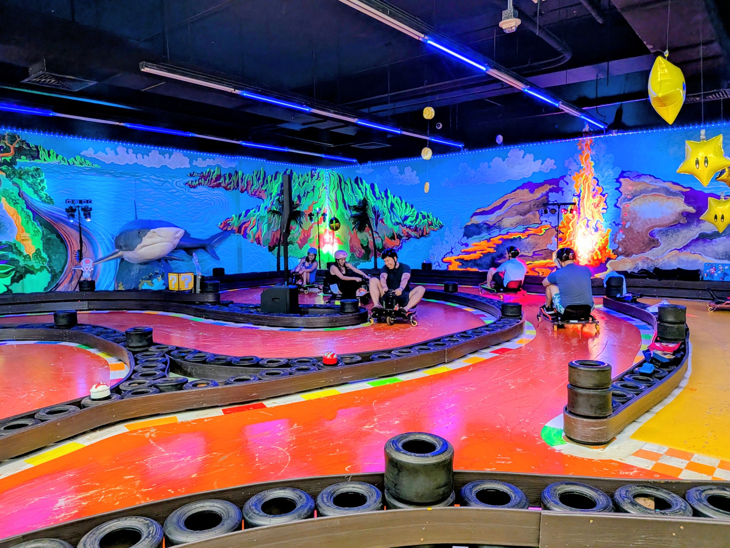 Adults drifting karts indoors in a cosmic blacklight environment with a vibrant lit wall mural