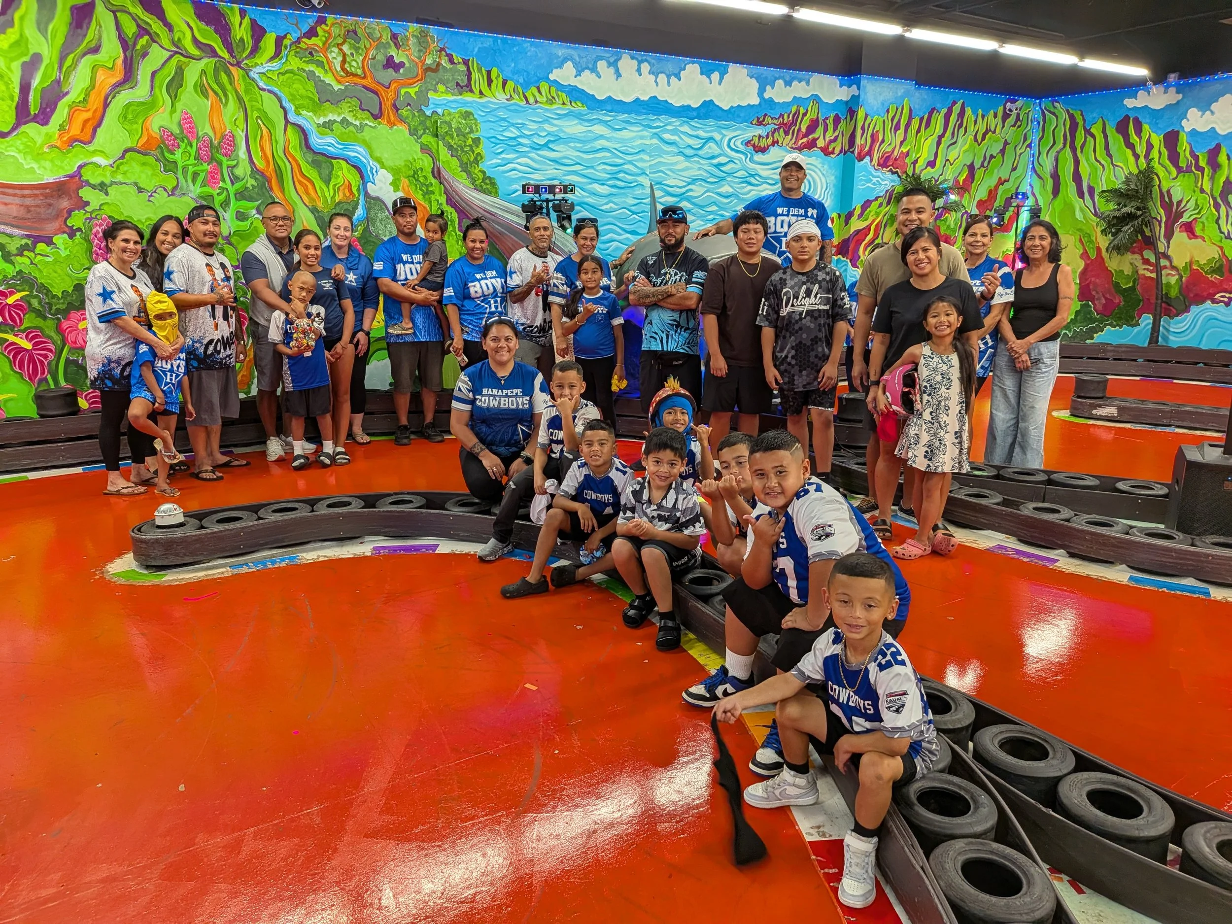 A group of children and adults pose inside a room with a colorful mural of a nature scene, including trees, mountains, and a river, painted on the walls. Some children are wearing sports jerseys, and everyone is smiling.