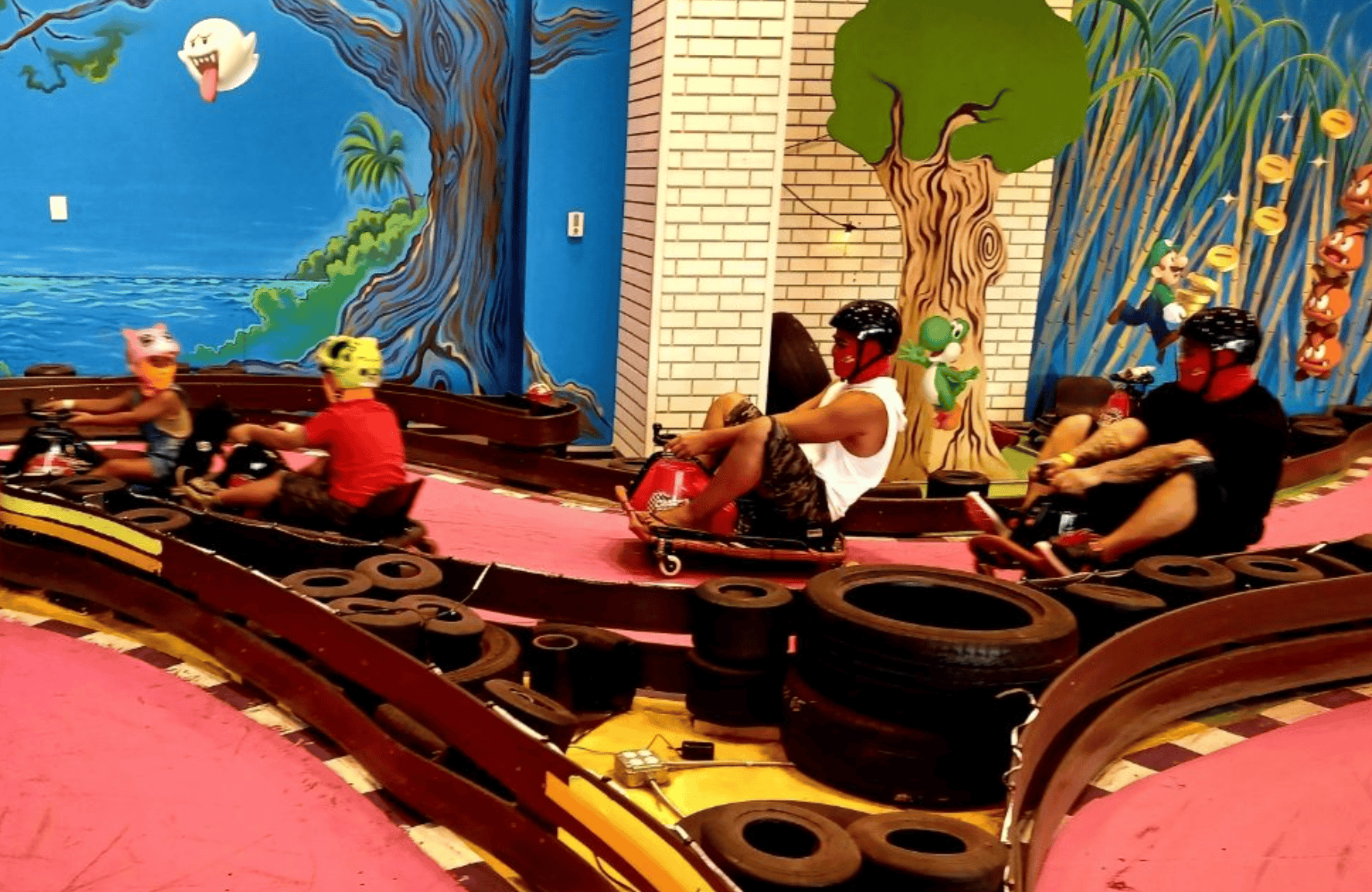 People riding small go-karts inside an indoor amusement center with colorful cartoon-themed murals of trees, animals, and a ghost on the walls.