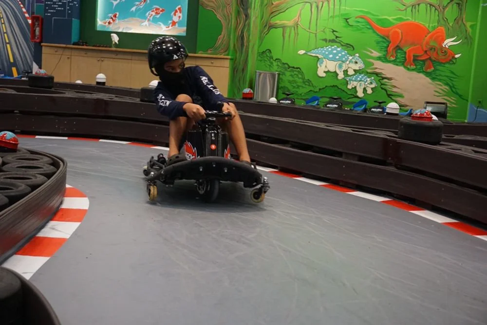 A young boy wearing a helmet and a face mask drifts a go-kart on an indoor track with colorful jungle mural walls.