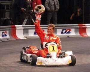 A race car driver holding a trophy and wearing a red racing suit, celebrating on a go-kart track with people in the background.