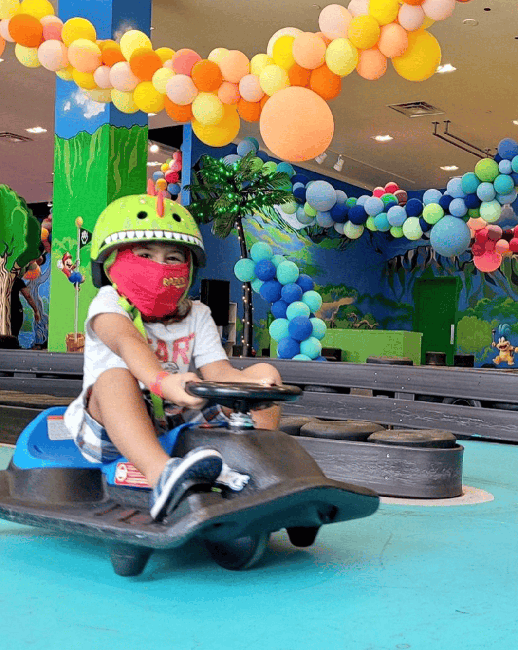 Child riding a smaller go-kart at an indoor amusement center with colorful balloon decorations and cartoon-themed murals.