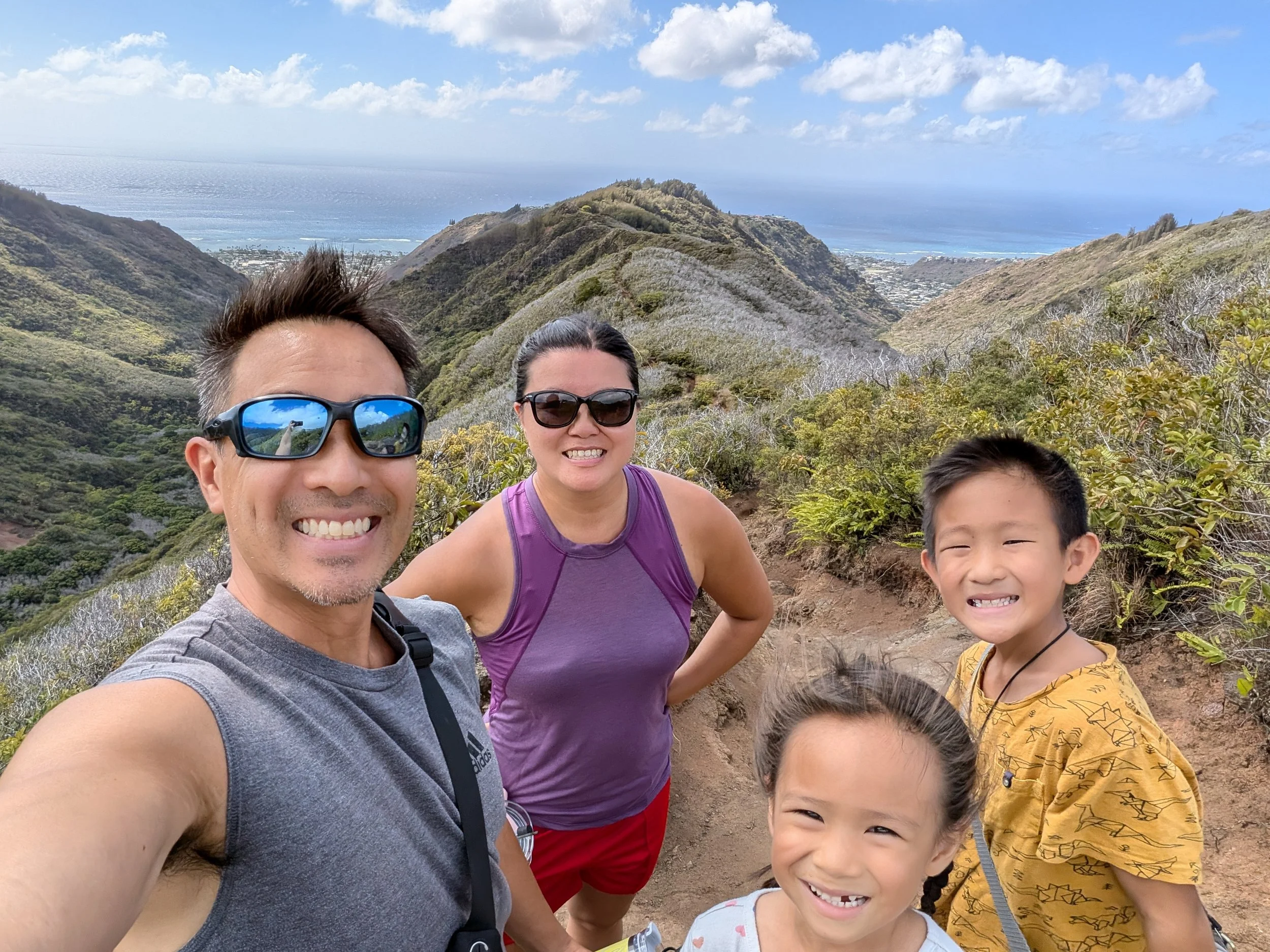 A family of four taking a selfie on a mountain trail with lush green hills and the ocean in the background. The man is wearing sunglasses and a gray sleeveless shirt, the woman is in purple, and the two children are smiling, one in a yellow shirt and the other in a white shirt with pink hearts.