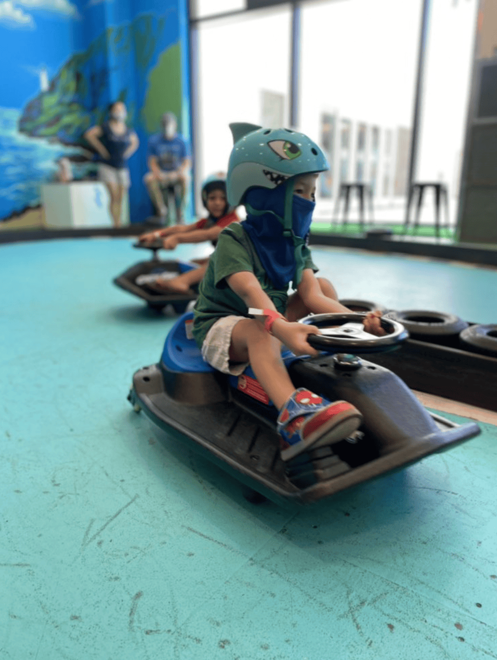 Children riding smaller go-karts indoors, with one child in the front wearing a helmet and face mask, and others behind him, in a play area with a blue floor and a mural on the wall.