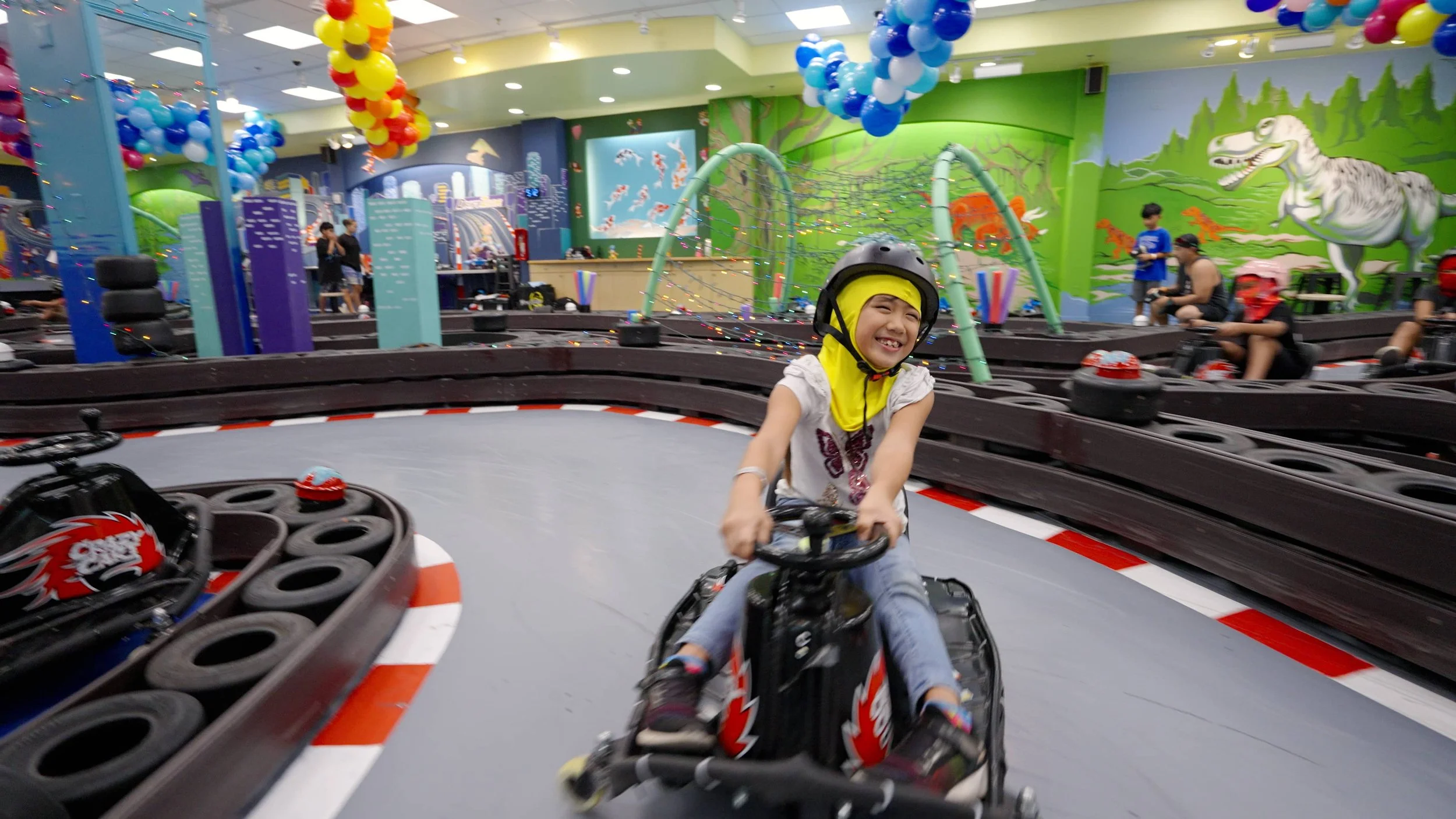 Children riding go-karts on an indoor go-kart track with colorful balloons and vibrant wall murals.