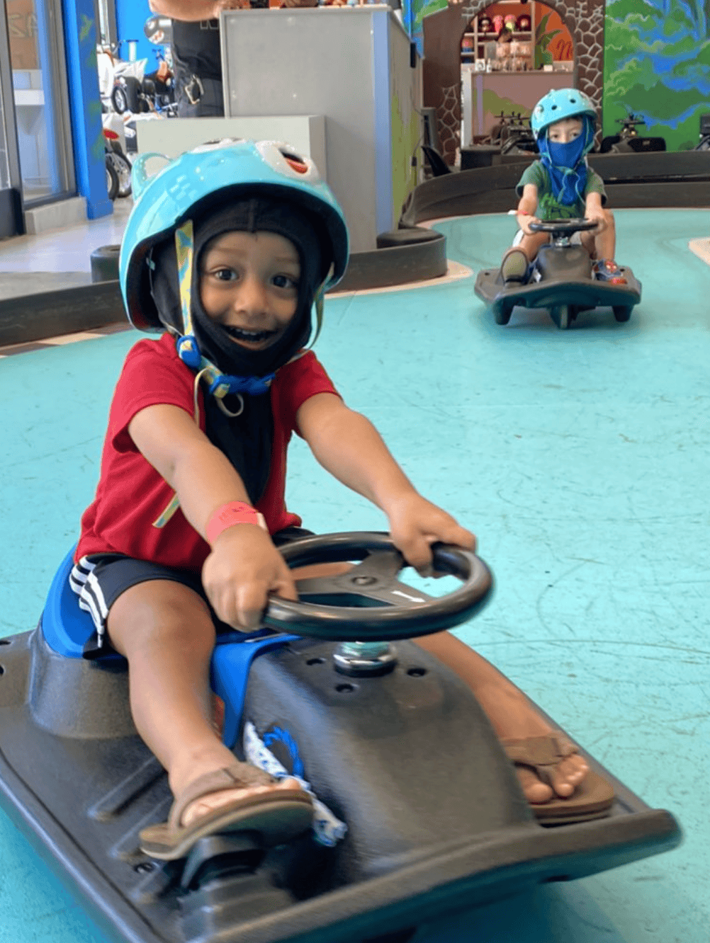 Two children riding smaller go-karts indoors, both wearing helmets and masks. The child in front is smiling, wearing a red shirt and black shorts, and the child in the background is sitting on a go-kart wearing a green shirt.