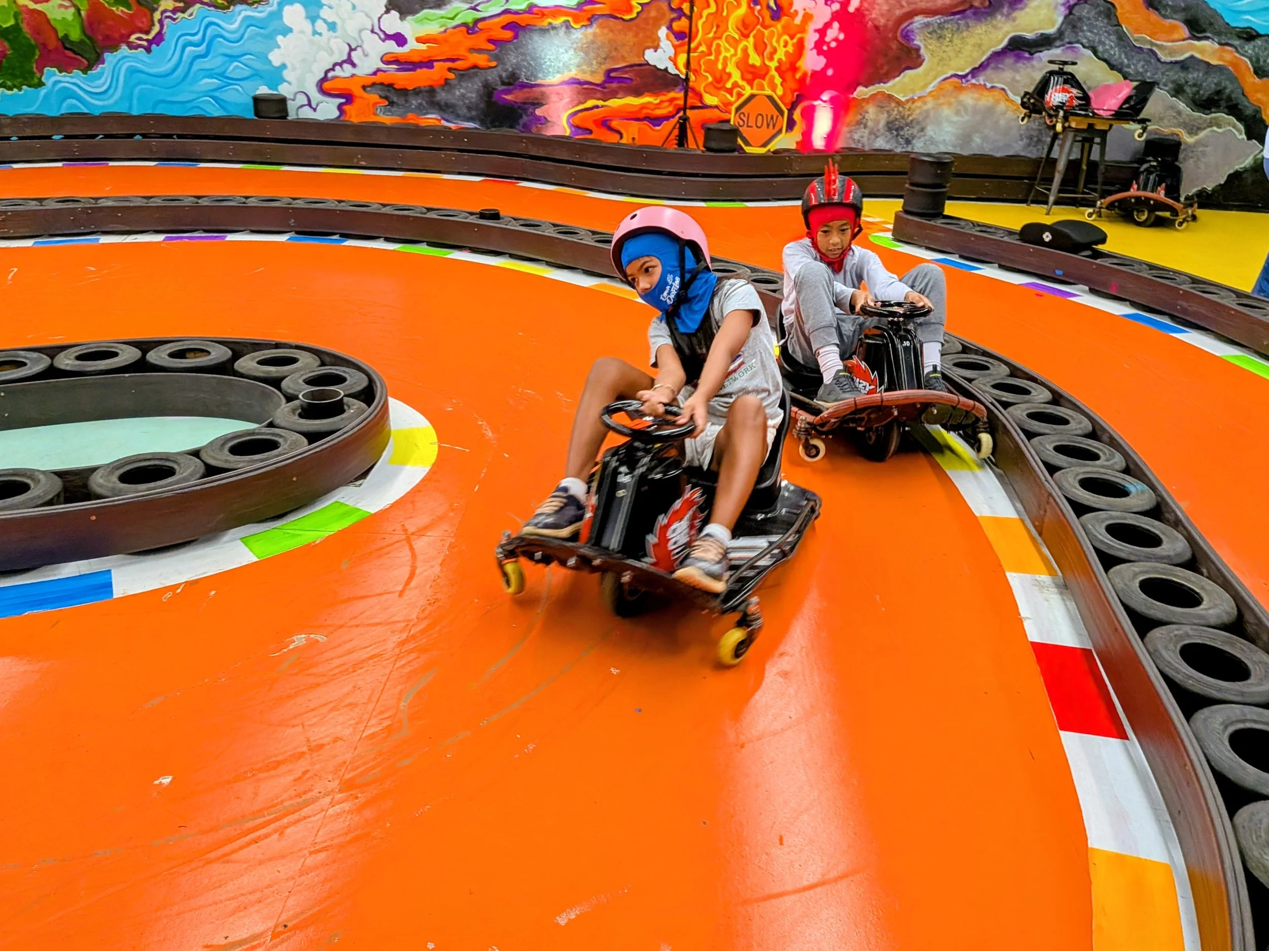 Two children wearing helmets are riding go-karts on an indoor race track with colorful, graffiti-style mural art in the background.