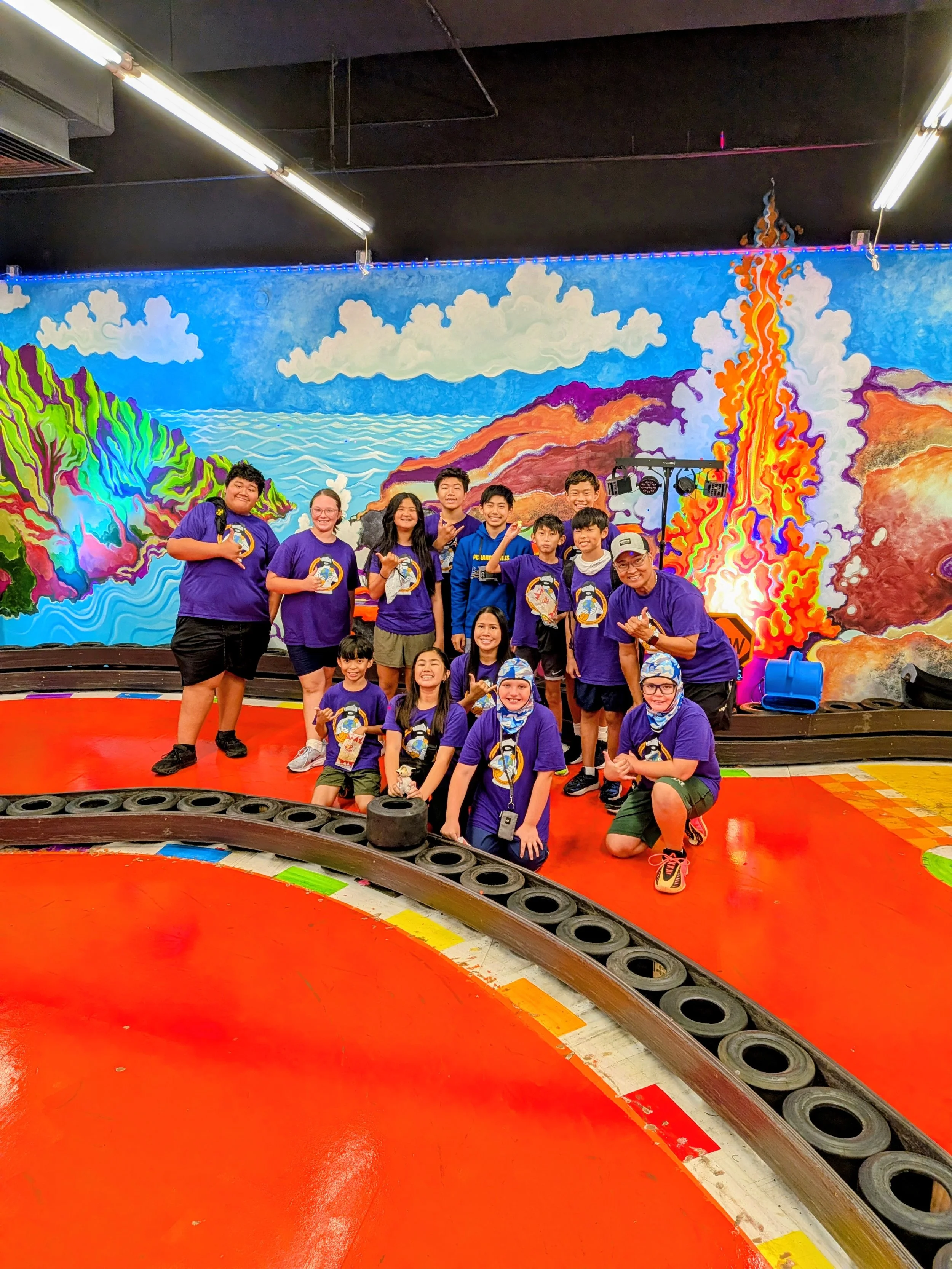 A group of children and adults in purple shirts posing for a photo at an indoor go-kart racing track with a bright, colorful mural of volcanic eruption in the background.