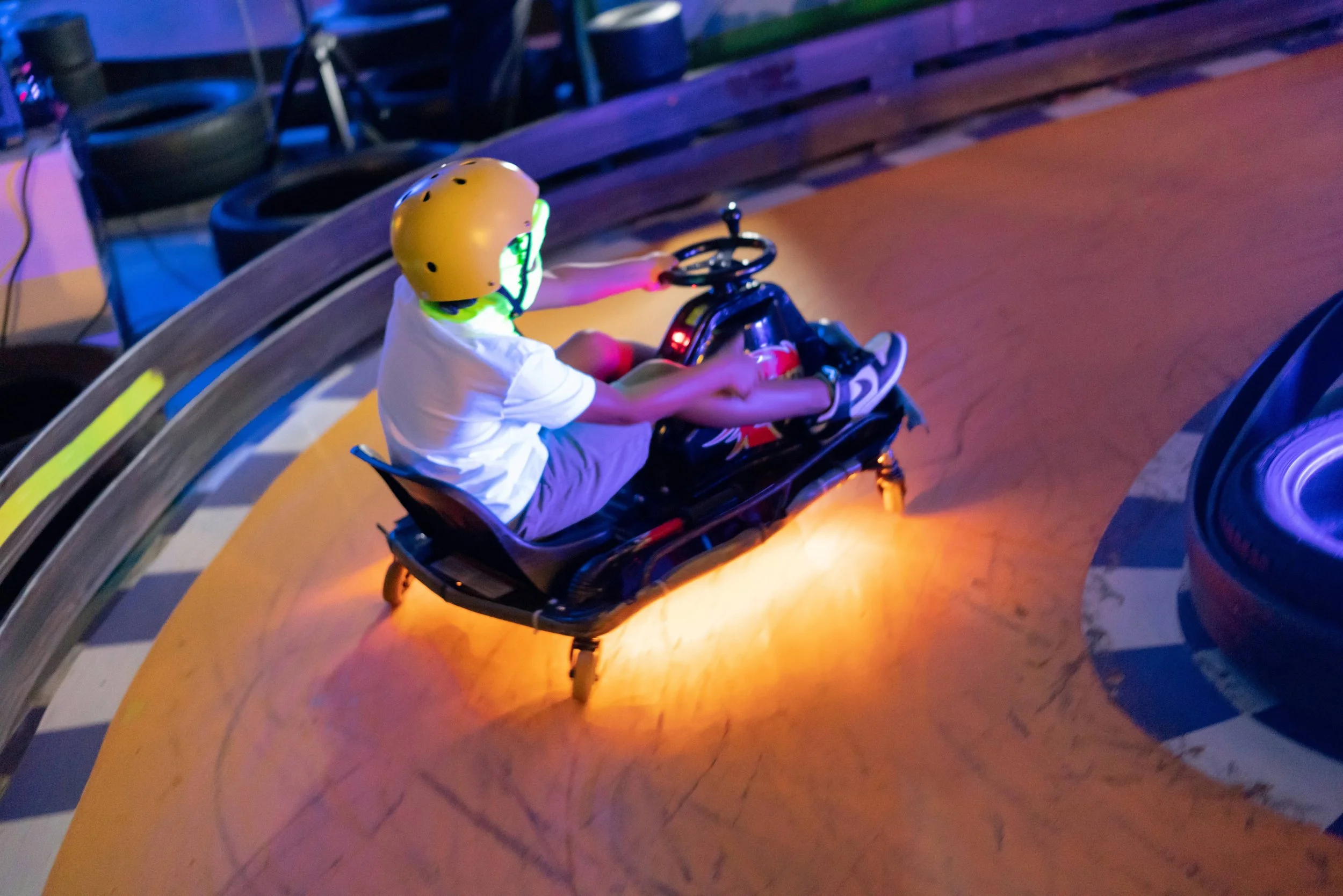 Child wearing a yellow helmet driving a go-kart indoors in a cosmic lit setting.
