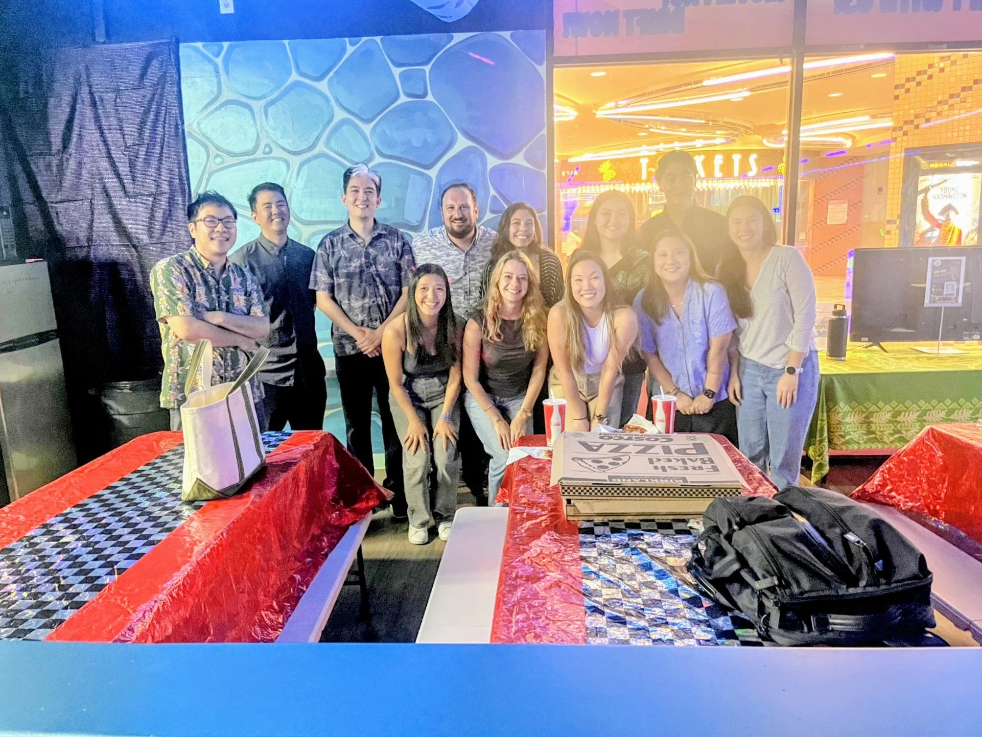 Group of people posing for a photo at a neon-lit entertainment venue, with tables and pizza boxes in the foreground.