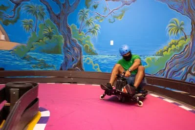 Person wearing a helmet and green shirt riding a go-kart on a pink track with a painted jungle mural in the background.