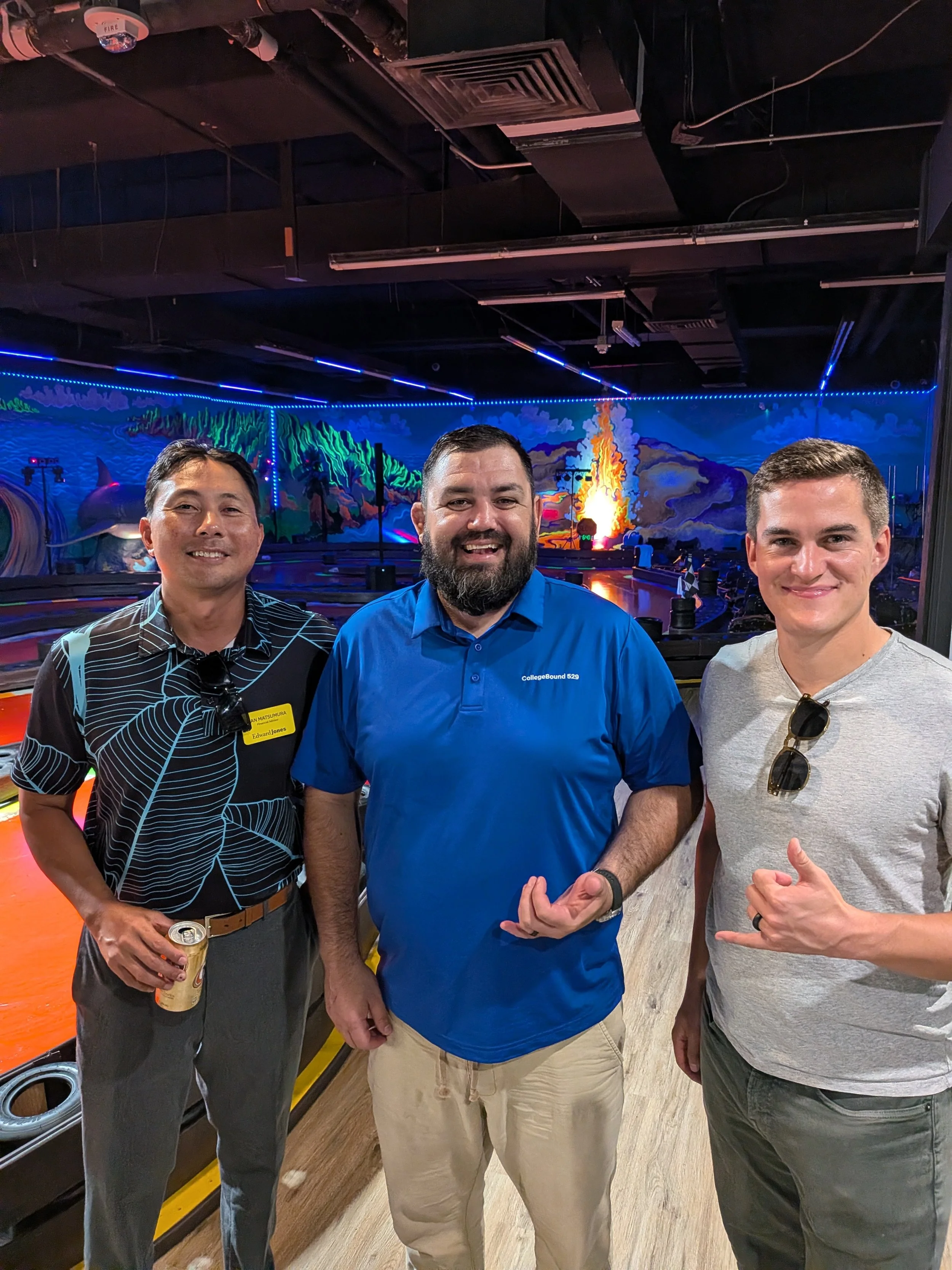 Three men posing and smiling at an amusement center, two of them making hand gestures, with a mural and race track in the background.