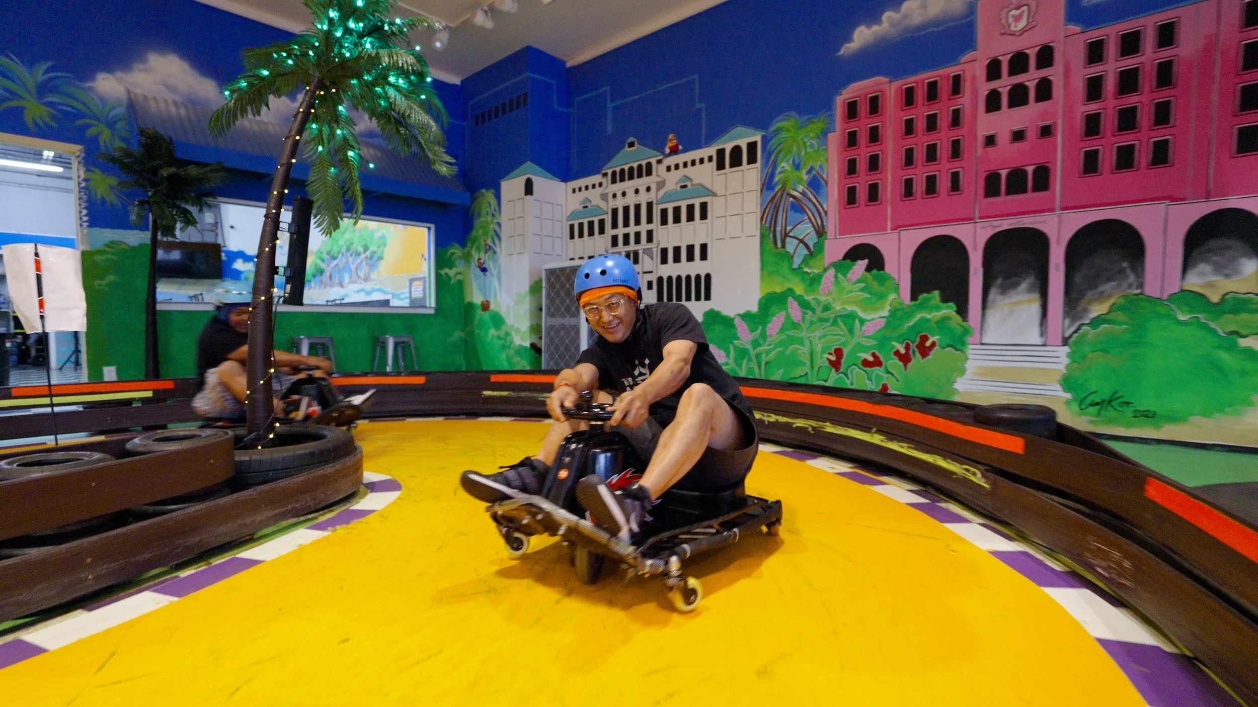 Person wearing a blue helmet and glasses driving a go-kart on an indoor track with colorful murals of buildings and palm trees in the background.