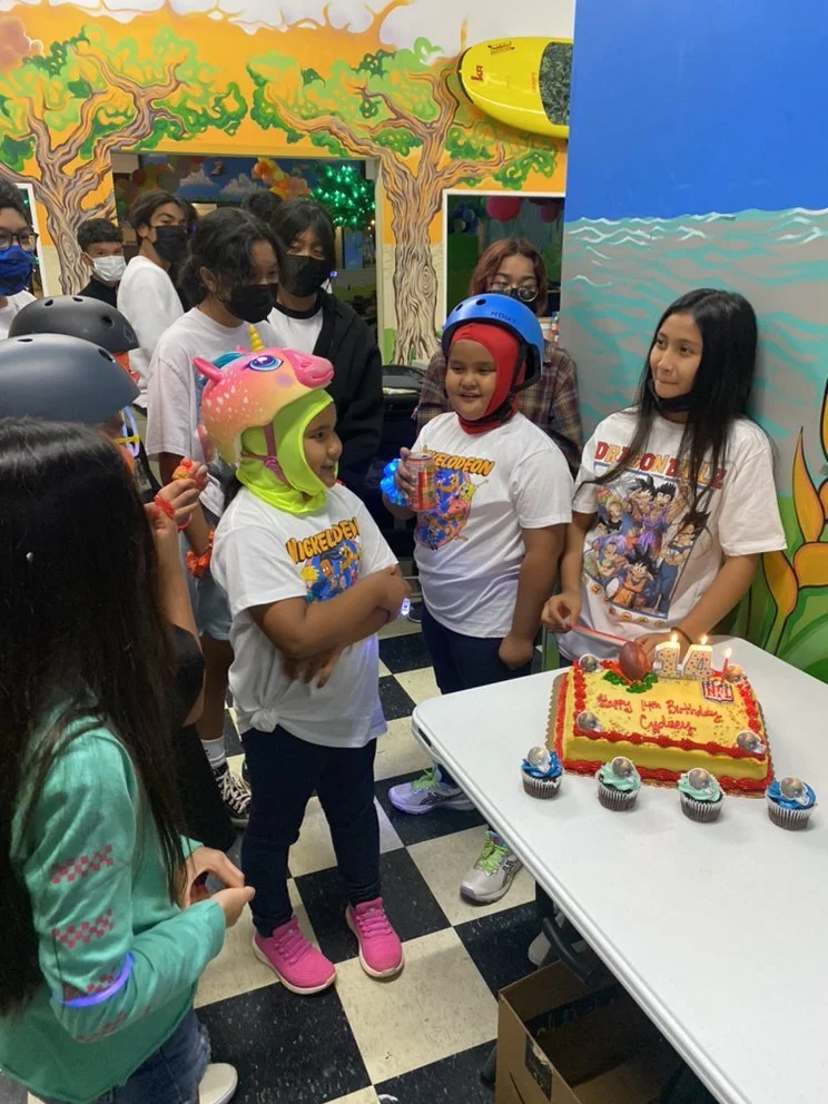 Children celebrating a birthday with a cake and cupcakes in an entertainment center decorated with colorful murals and a surfboard.