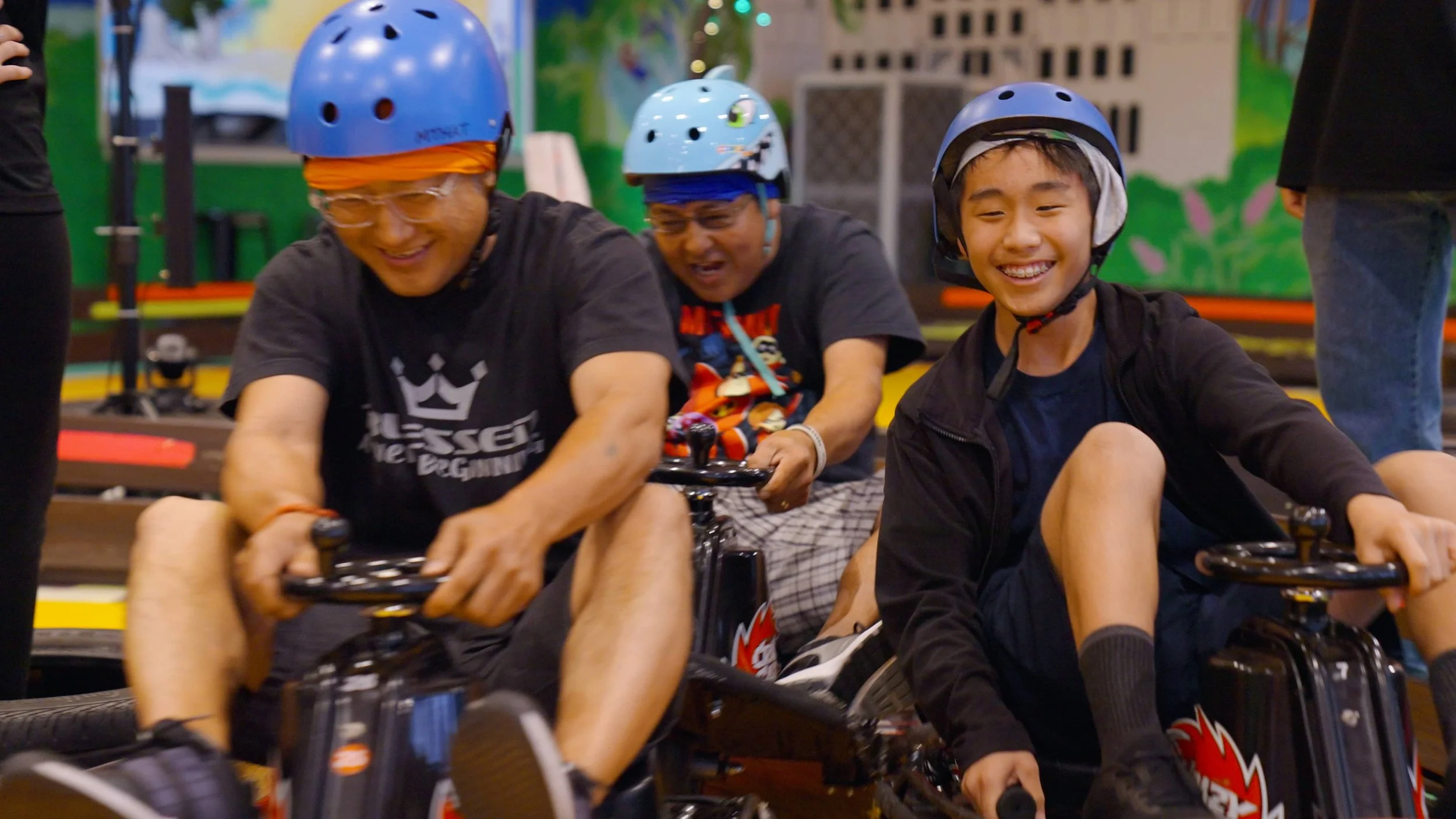 Three people of different ages wearing helmets and smiling while driving electric go-karts at an indoor go-kart track.
