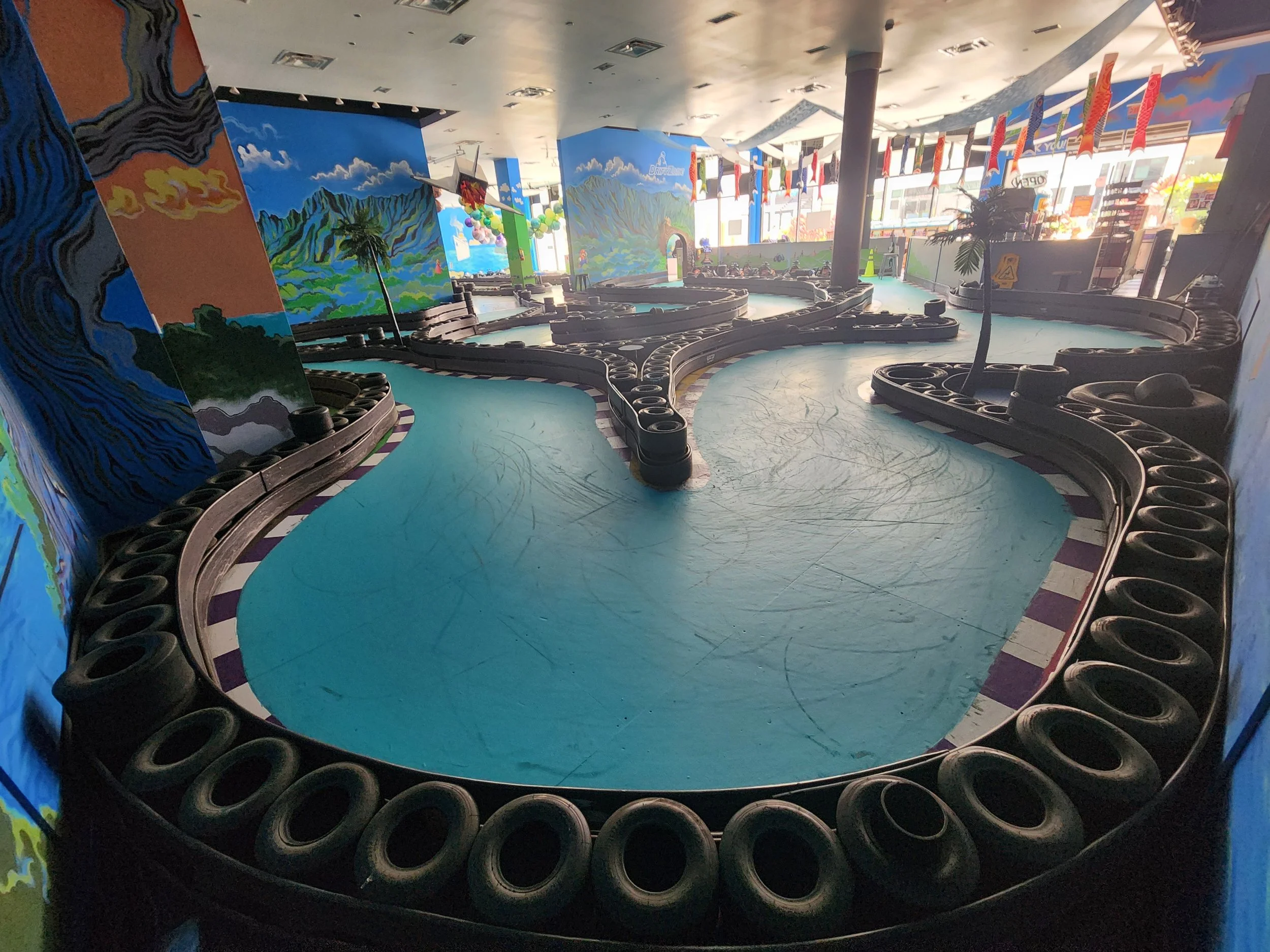 Indoor mini race track with winding lanes surrounded by rubber tires, colorful mural walls, and large windows with sunlight streaming in.