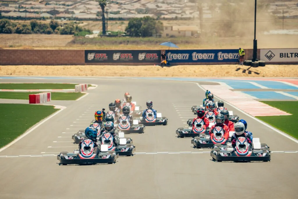 Go-kart racers lined up on the starting grid at a racetrack, ready for the race to begin, under a sunny sky.
