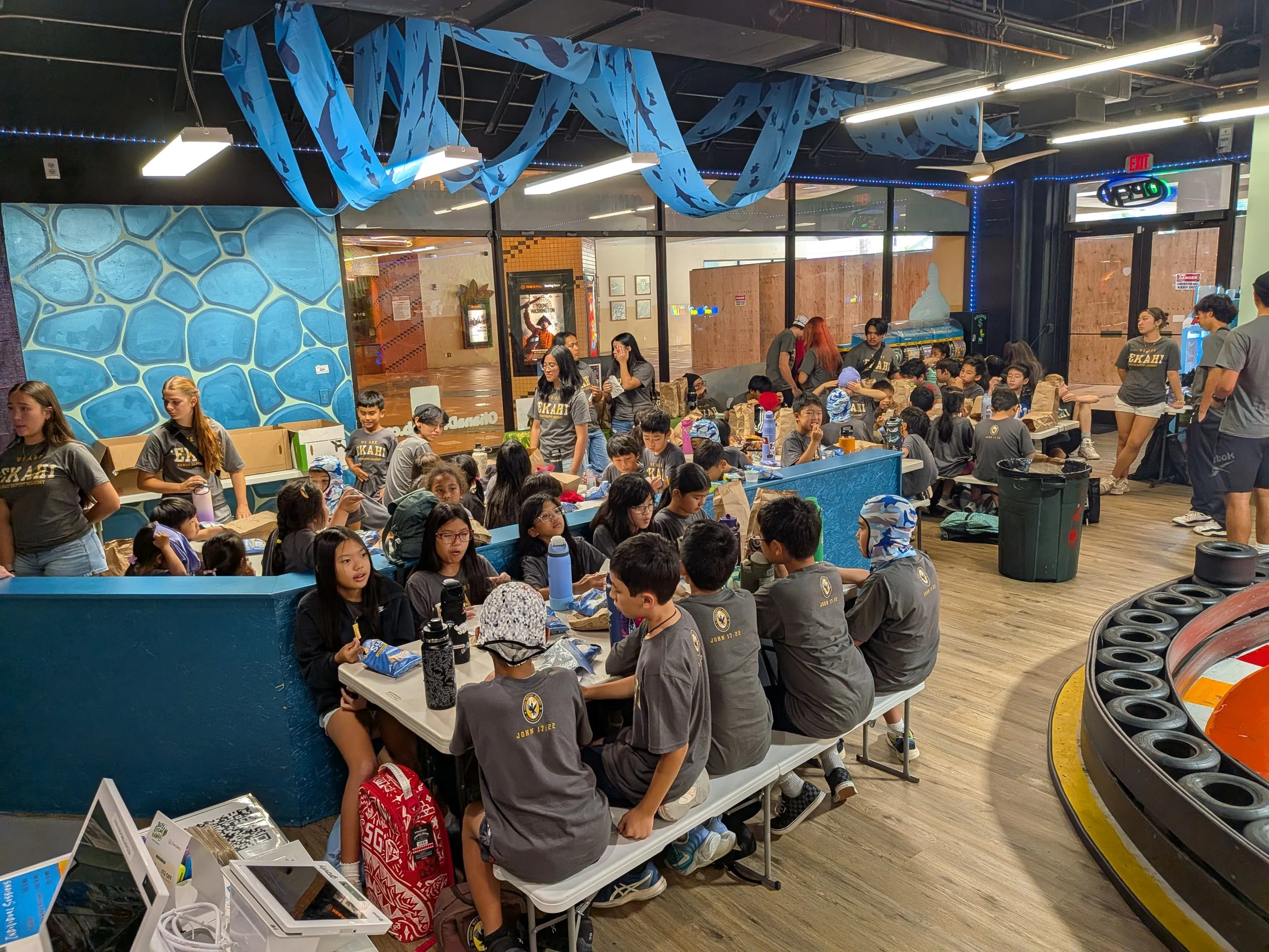 A large indoor room filled with children and adults participating in an event. Many children are seated at long tables with paper bags, and adults are standing or sitting among them. Blue paper streamers hang from the ceiling, and there are various p