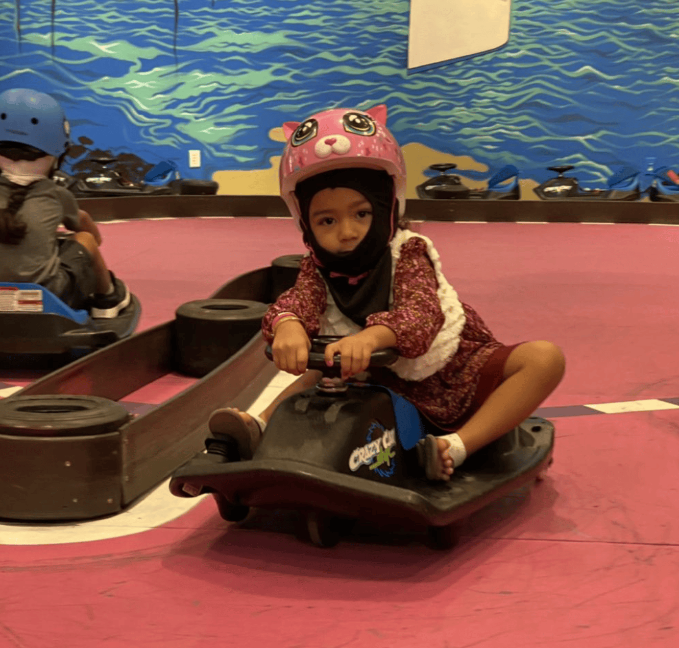 A young girl wearing a pink helmet with animal face design, a patterned long-sleeve shirt, and a white vest, is sitting on a go-kart on a pink track with a blue wave mural in the background.