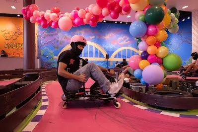Person drifting a go-kart sideways on an indoor track decorated with colorful balloons and a rainbow backdrop.
