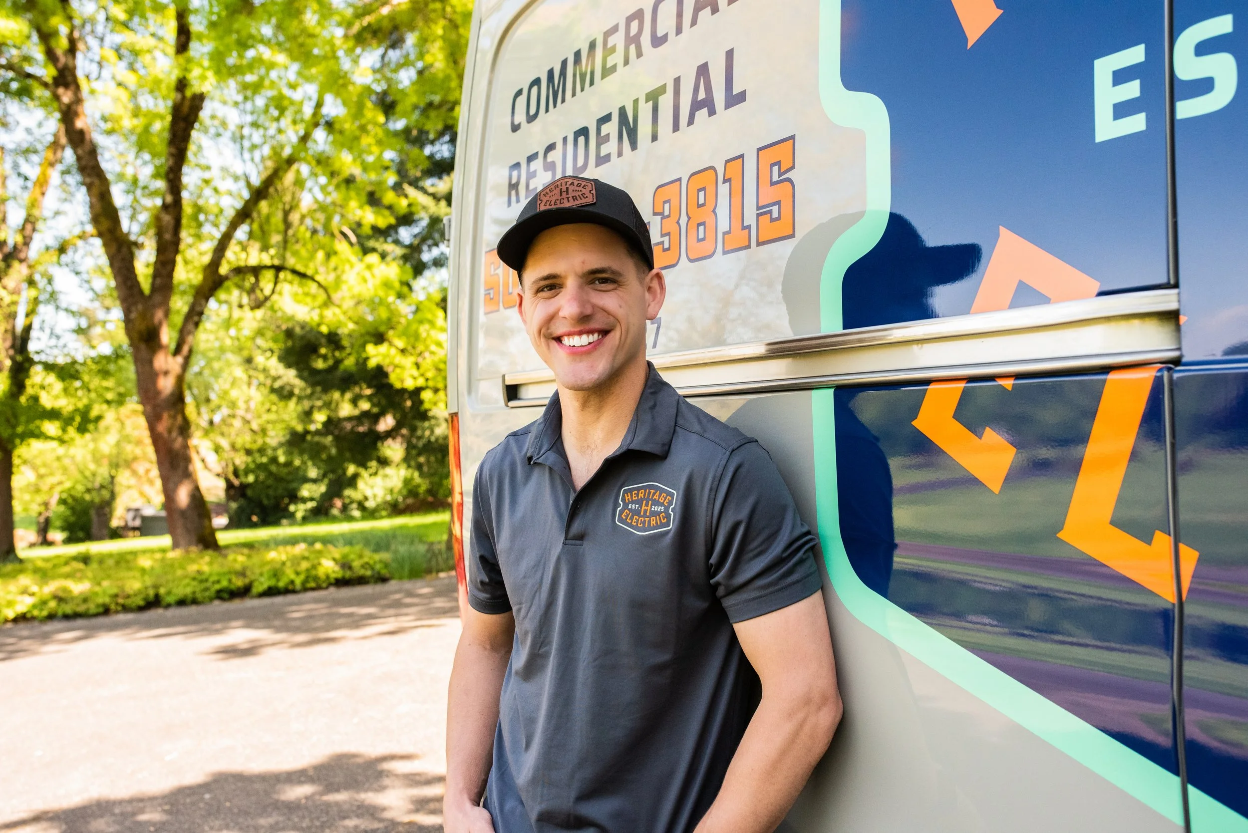 A smiling man in a black baseball cap and a gray uniform stands outdoors next to a colorful service vehicle with the words "Commercial Residential" and a phone number on it. Green trees with sunlight filtering through are in the background.