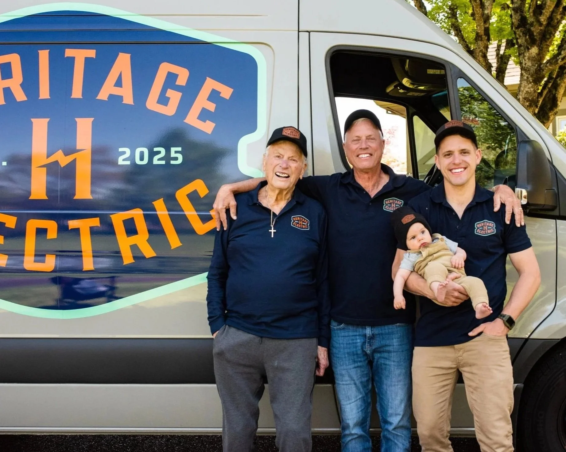 Group of four people, two men, one woman, and a baby, standing in front of a vehicle with a logo that says 'Heritage Electric'. The group is smiling and appears to be happy, with trees and a house in the background.