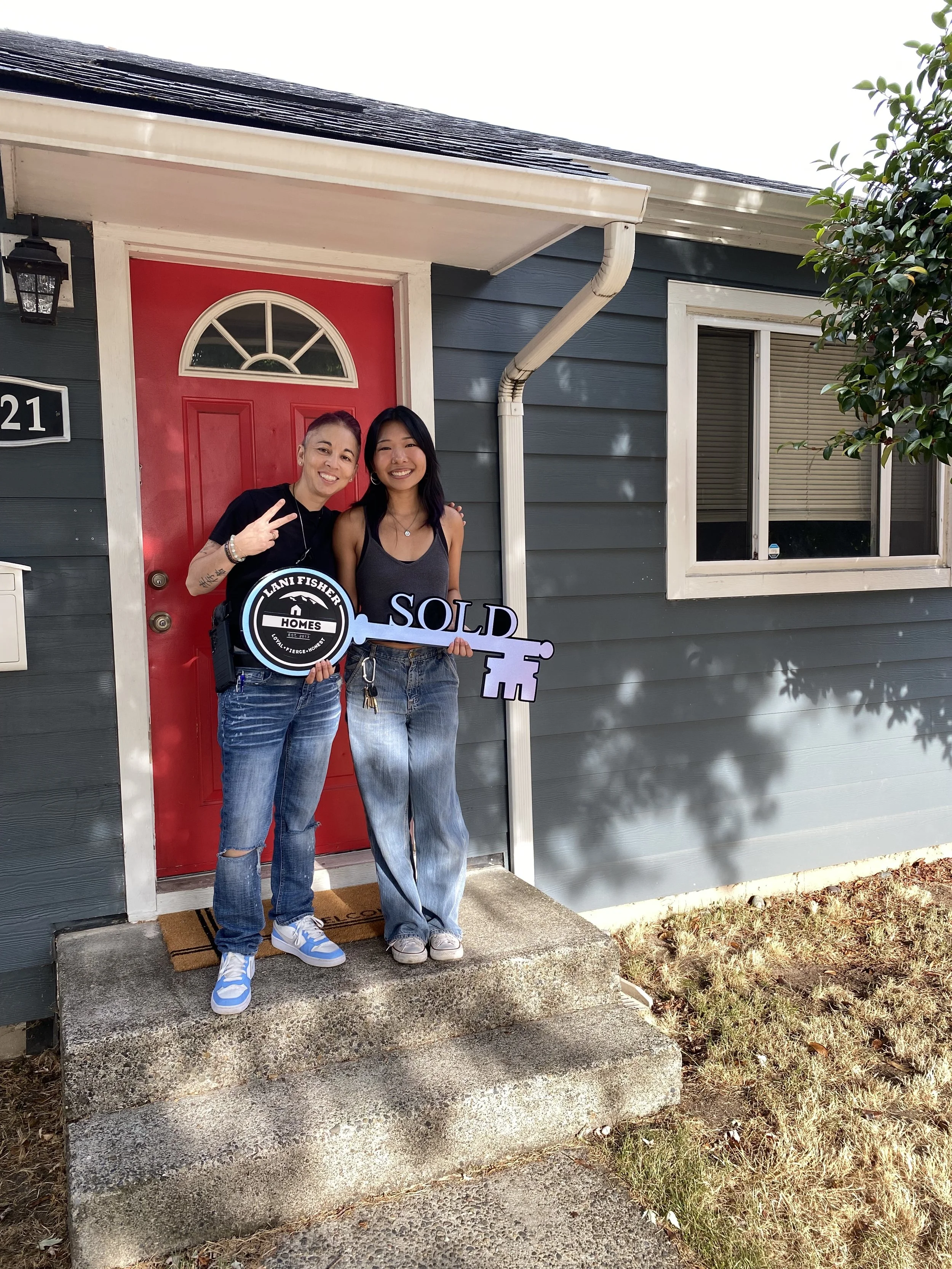 Pacific Northwest Realtor and U.S. Veteran Lani Fisher celebrating with a first-time home buyer in her early 20s after closing on her new home — standing on the front steps with a large “SOLD” key featuring the Lani Fisher Homes logo.