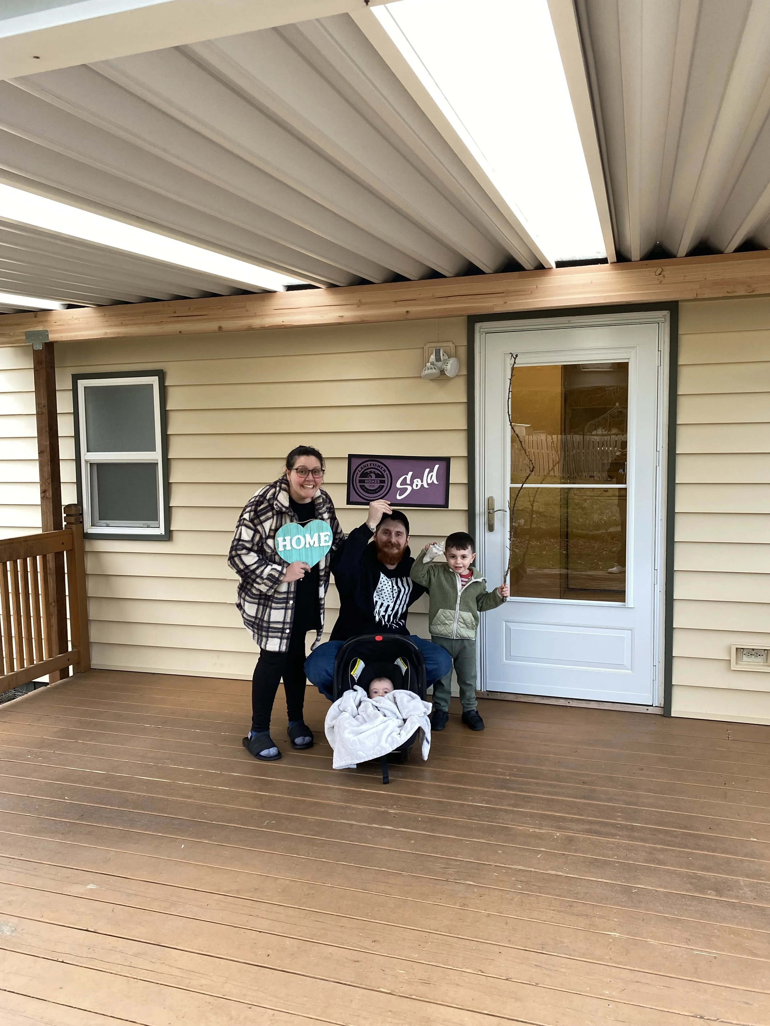 Young family celebrating buying their first home with Pacific Northwest Realtor and U.S. Veteran Lani Fisher — standing on the porch with “Home” and “Sold” signs after closing with Lani Fisher Homes, marking the start of a new chapter together.