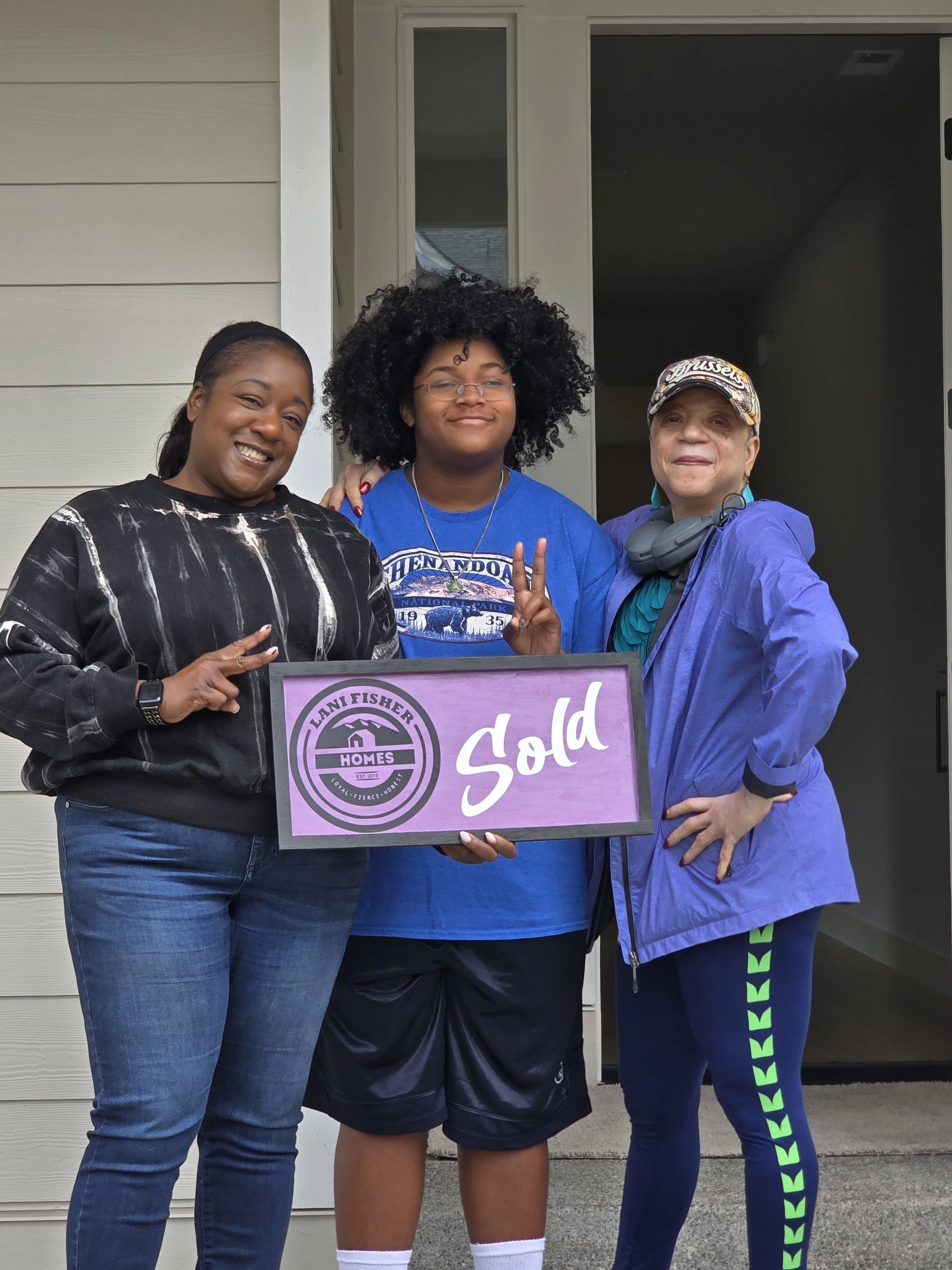 Three generations of women celebrating the sale of a family home with Pacific Northwest Realtor and U.S. Veteran Lani Fisher — holding a purple “Sold” sign outside their new home, representing family, legacy, and homeownership with Lani Fisher Homes.