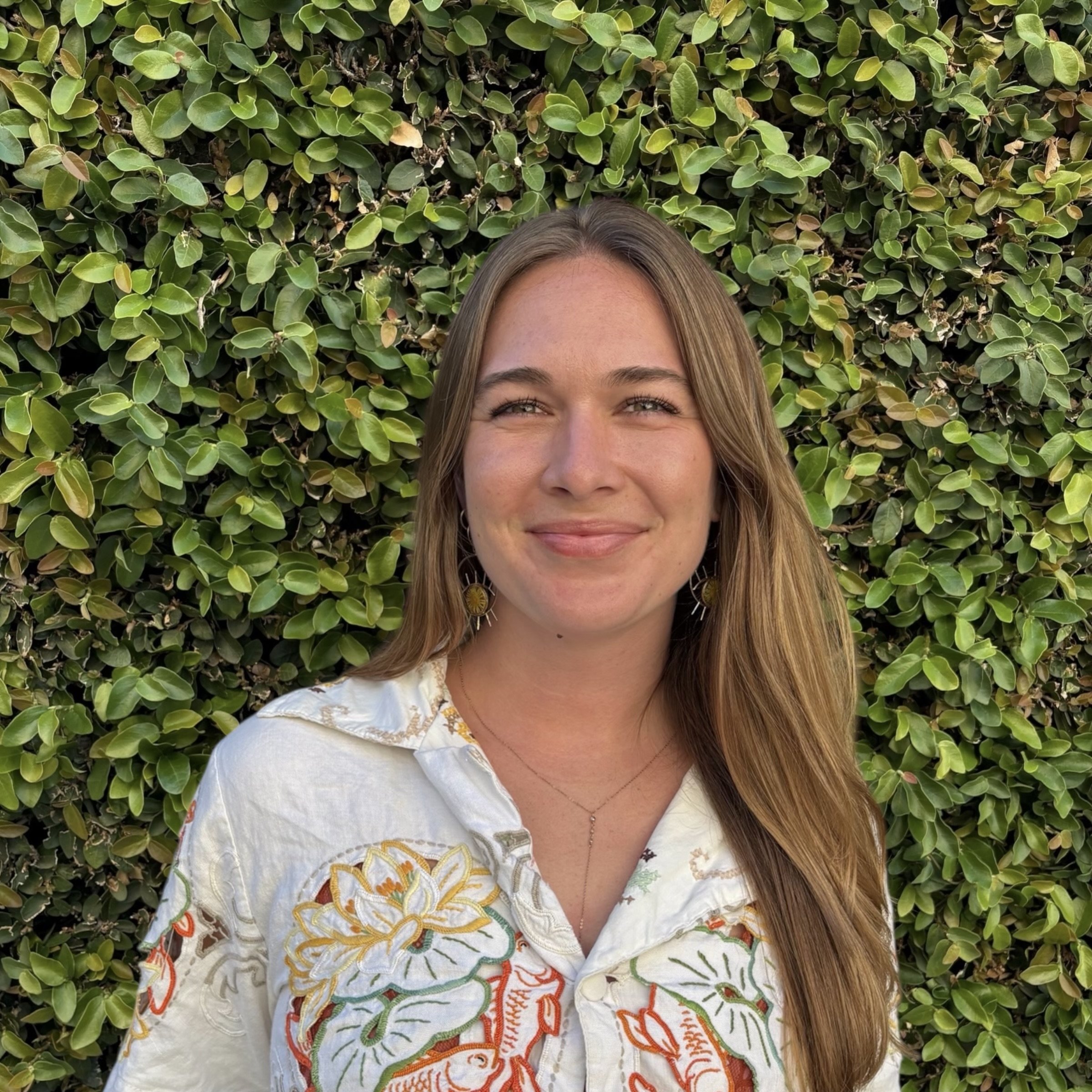 Young woman with long light brown hair smiling, standing in front of a green leafy hedge, wearing a white embroidered shirt and earrings.