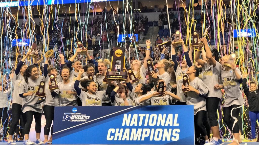 A group of female UCLA gymnasts celebrating their victory with trophies after winning the National Championship event, surrounded by colorful streamers and confetti.