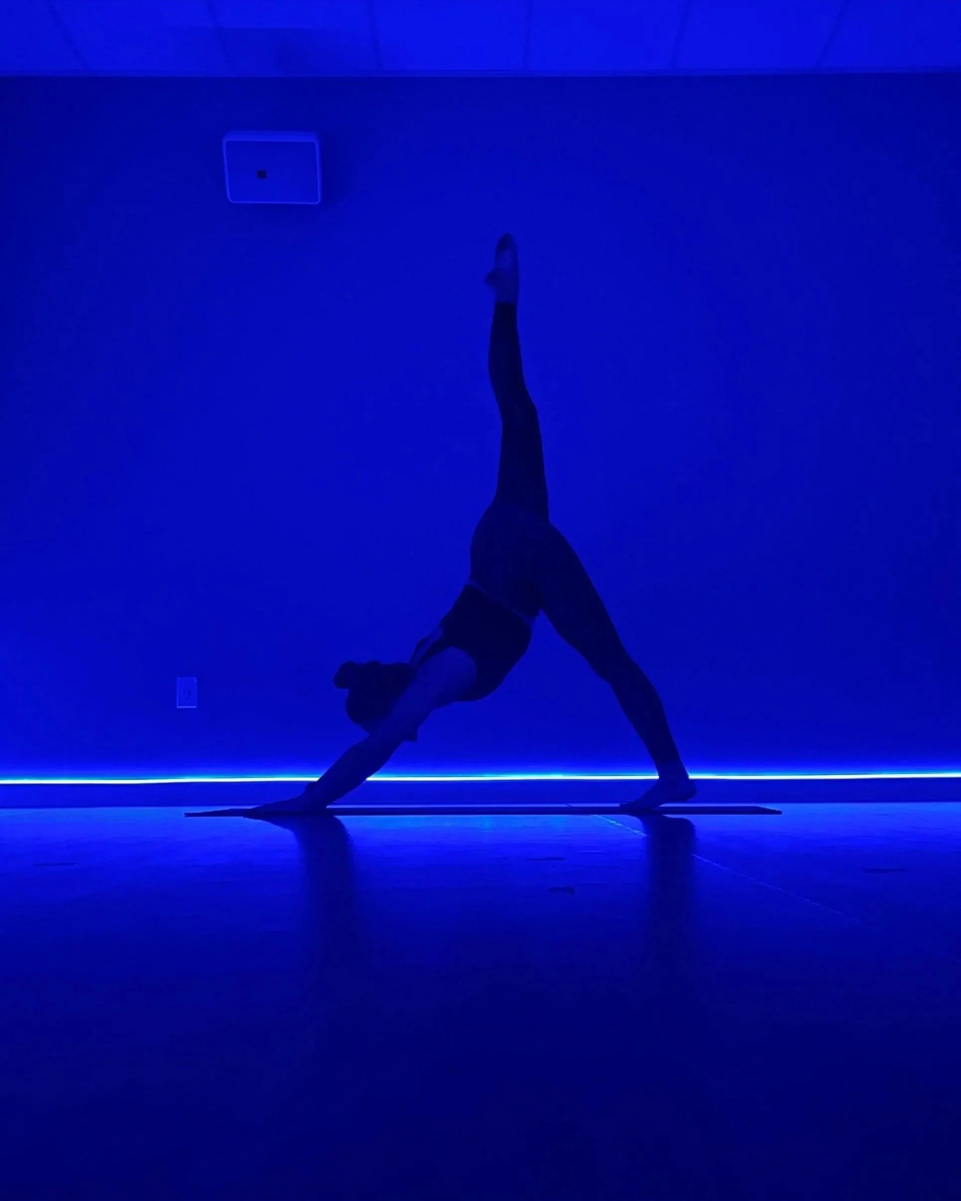 A person practicing yoga in a dimly lit room with blue lighting, performing a downward dog pose on a yoga mat.