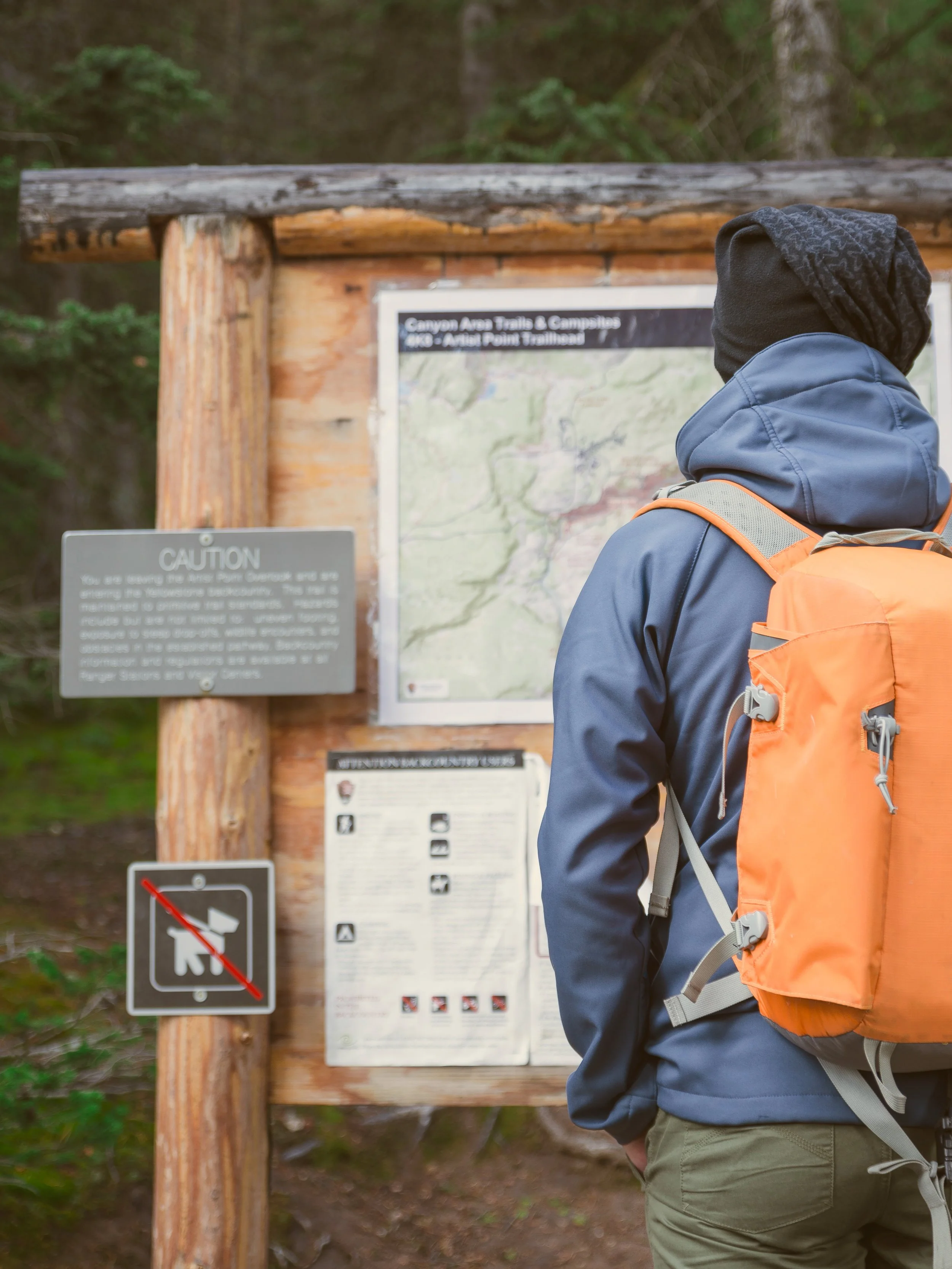 Creative signage services printing church design Hiker with orange backpack reading an information board at a trailhead in a wooded area.