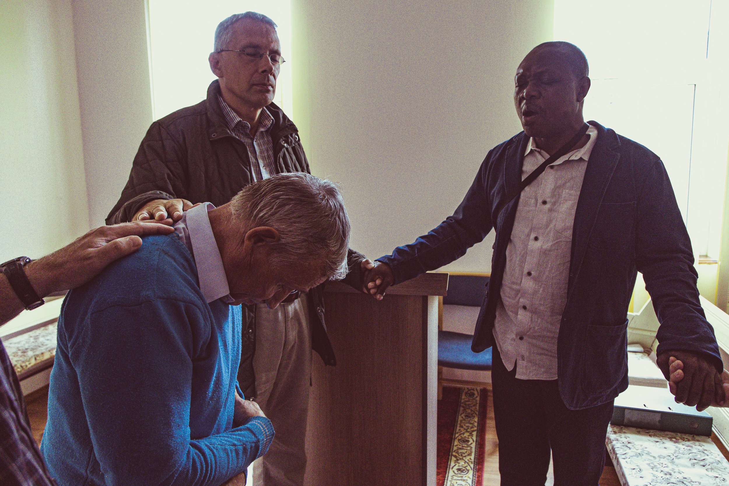 pastors, missions, missionaries, mission trip, mission help four men praying in a church, one man kneeling with head bowed, others with eyes closed, some with hands on his shoulders.
