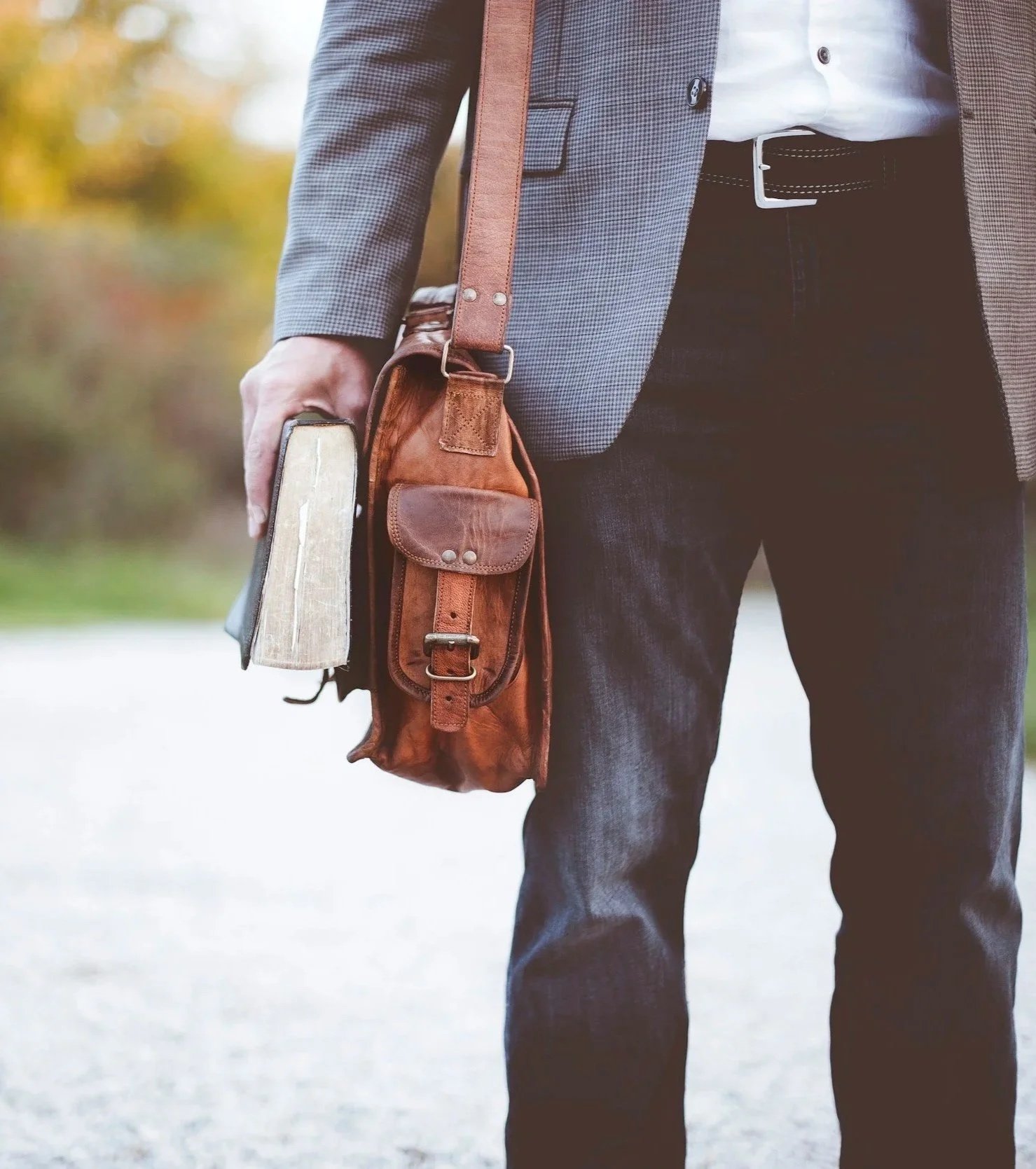 Authority of the Bible, Church, Consulting, Coaching, Pastor, Man holding a book and carrying a leather satchel, wearing a grey blazer and dark jeans, outdoors with autumn trees in the background.