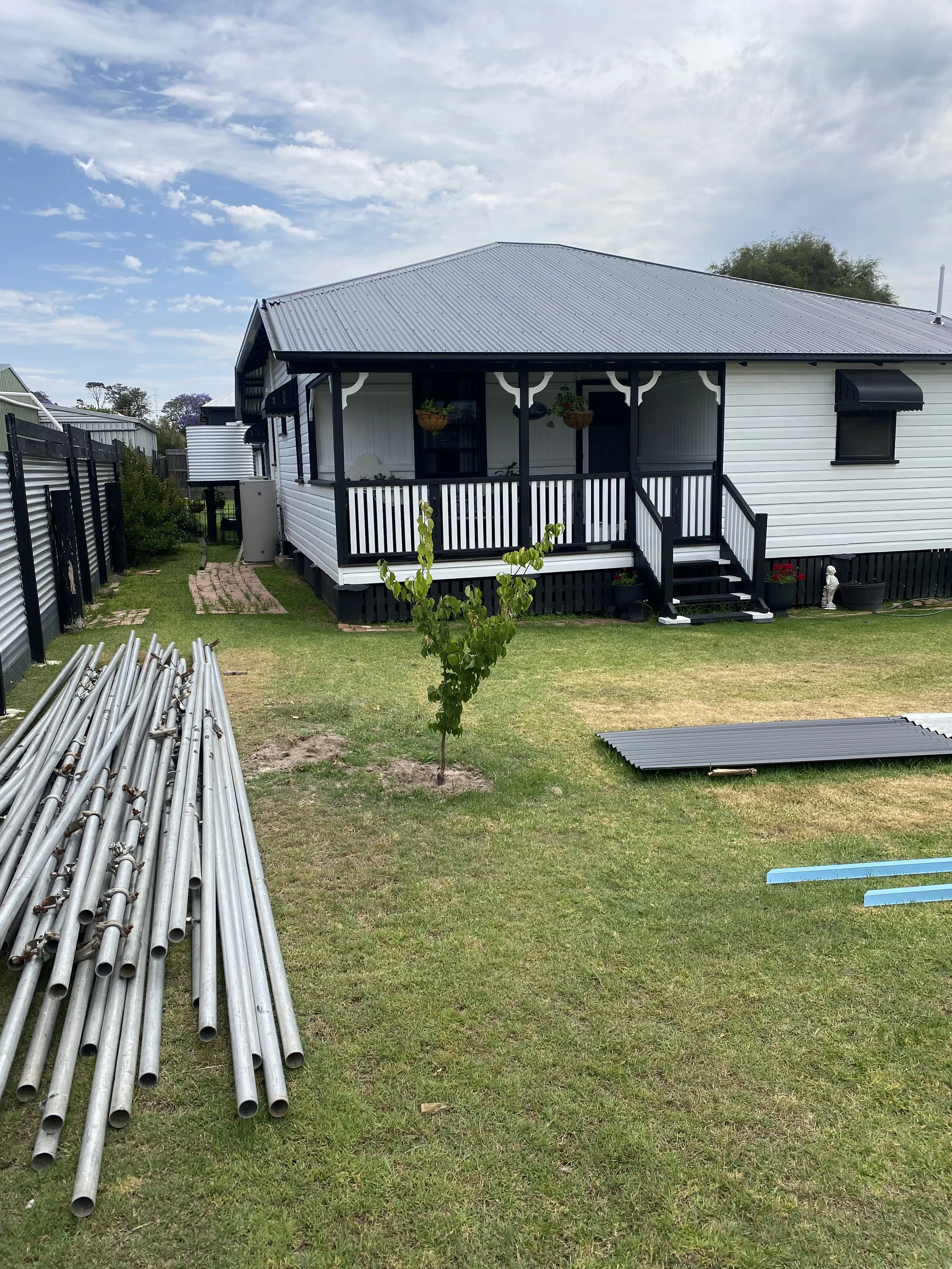 A backyard with a small tree, a black and white house with a porch, a pile of metal poles, and some construction materials on the grass.