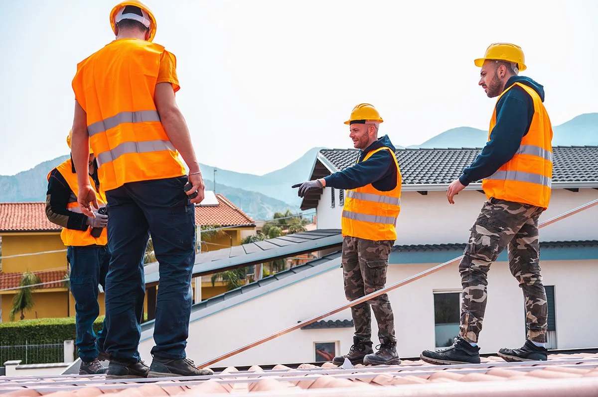 Construction workers wearing orange safety vests and yellow helmets on a roof, with houses and mountains in the background.