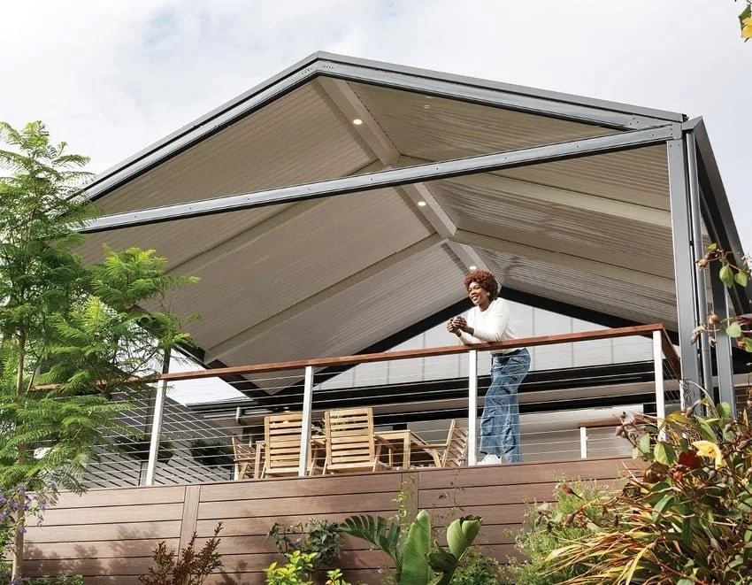 A woman standing on a balcony of a modern house with an open roof, holding a phone, surrounded by greenery.