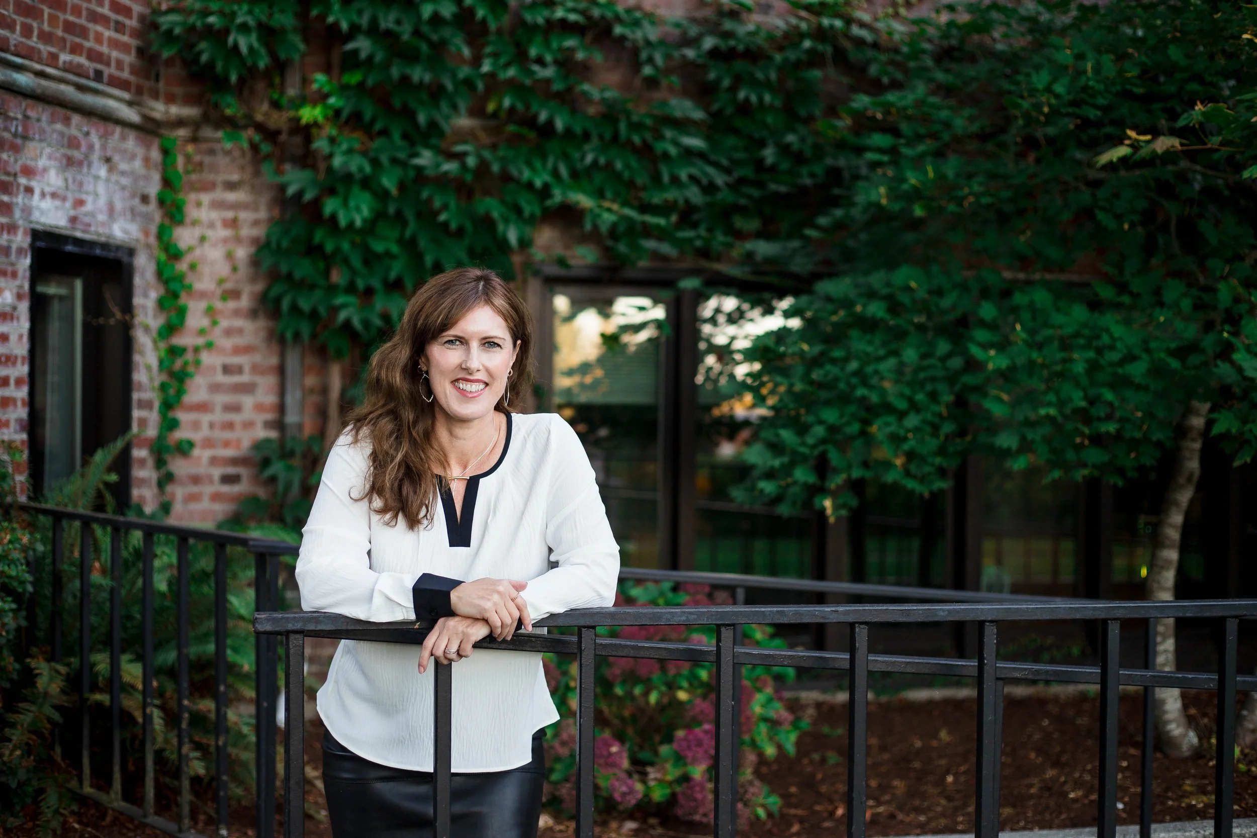 A woman with long brown hair, wearing a white blouse with black accents, standing outdoors behind a black metal railing, smiling at the camera with a garden and brick building in the background.