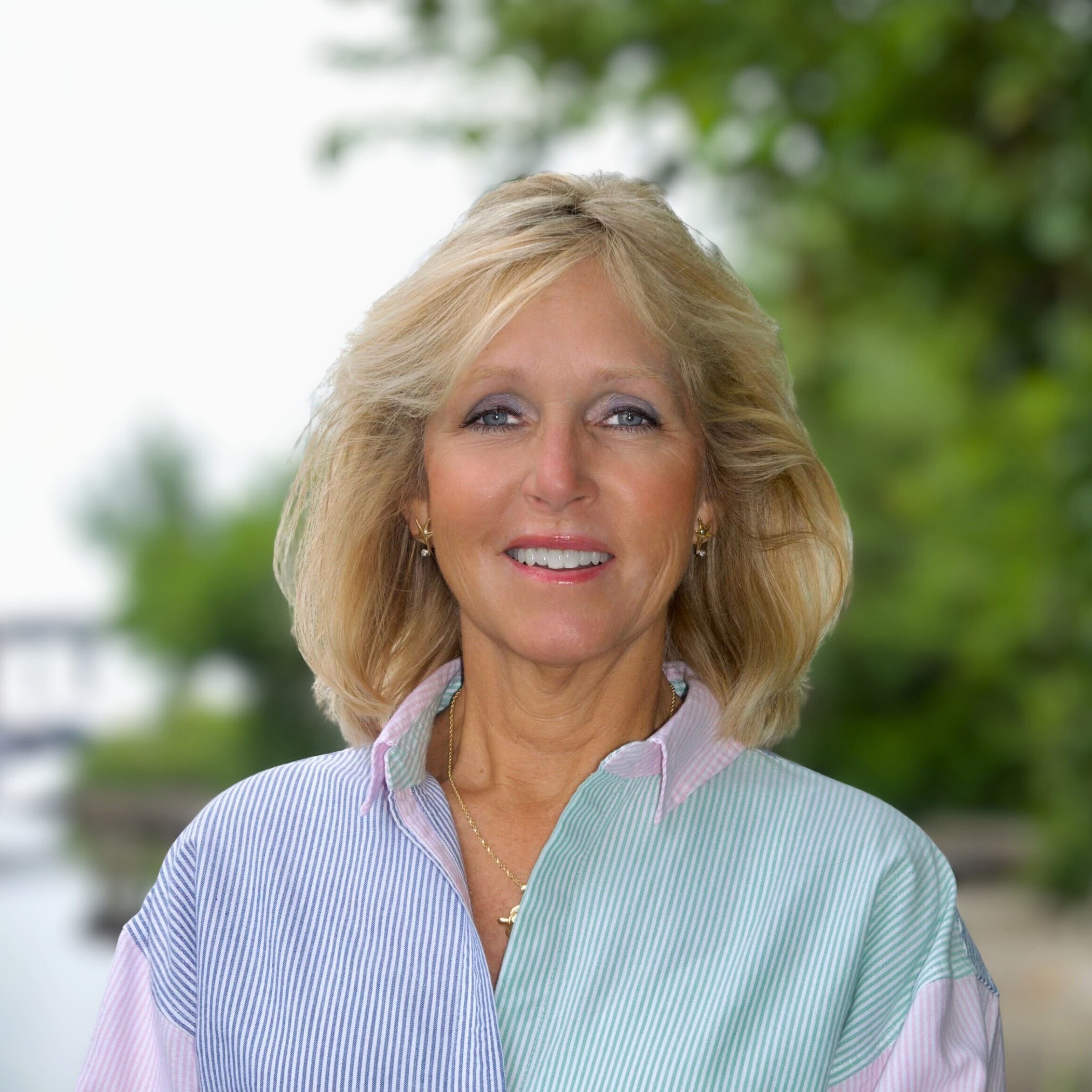 A woman with shoulder-length blonde hair, wearing a pastel-colored striped shirt, smiling outdoors with green trees blurred in the background.