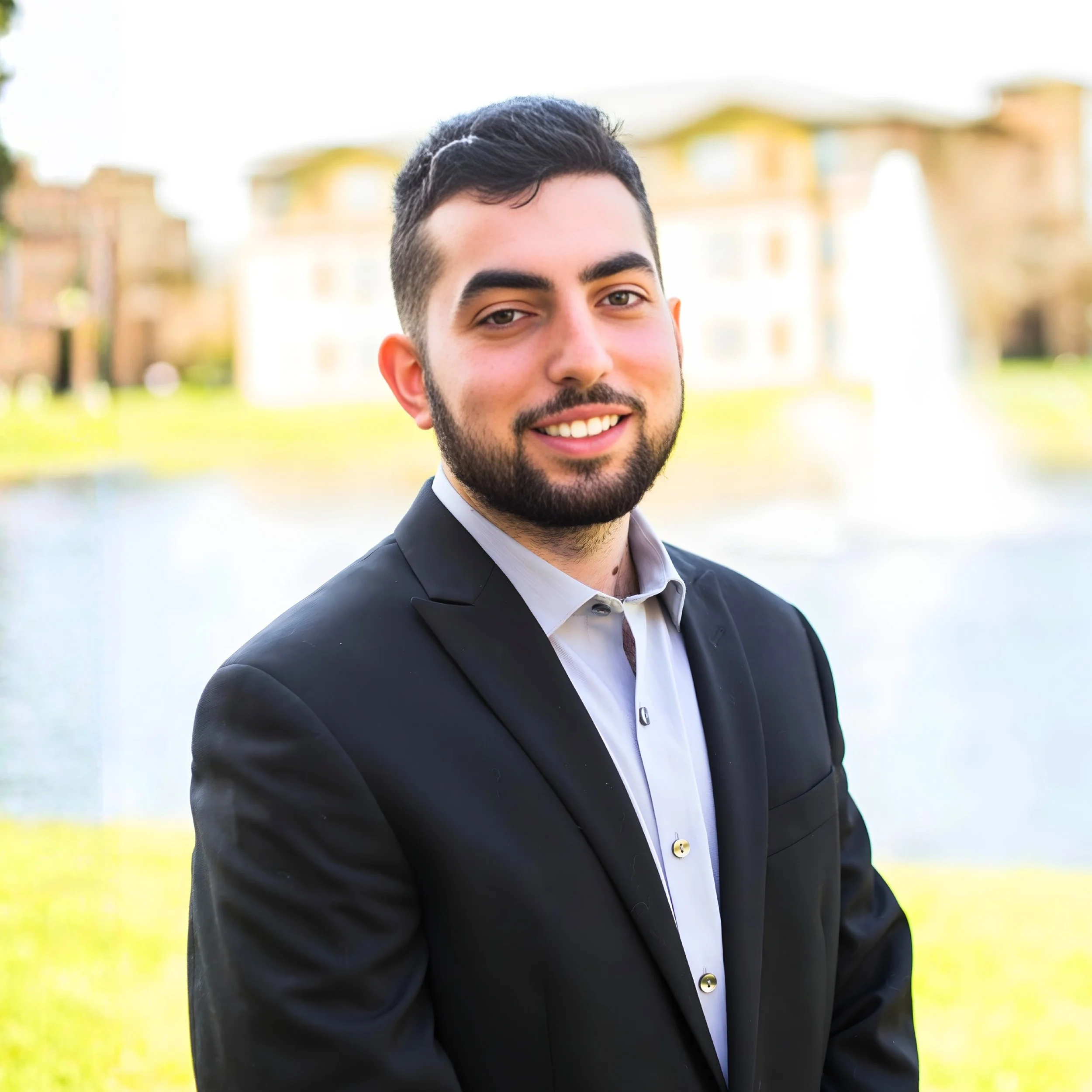 A young man in a black suit and white shirt smiling outdoors near a pond with a fountain and buildings in the background.